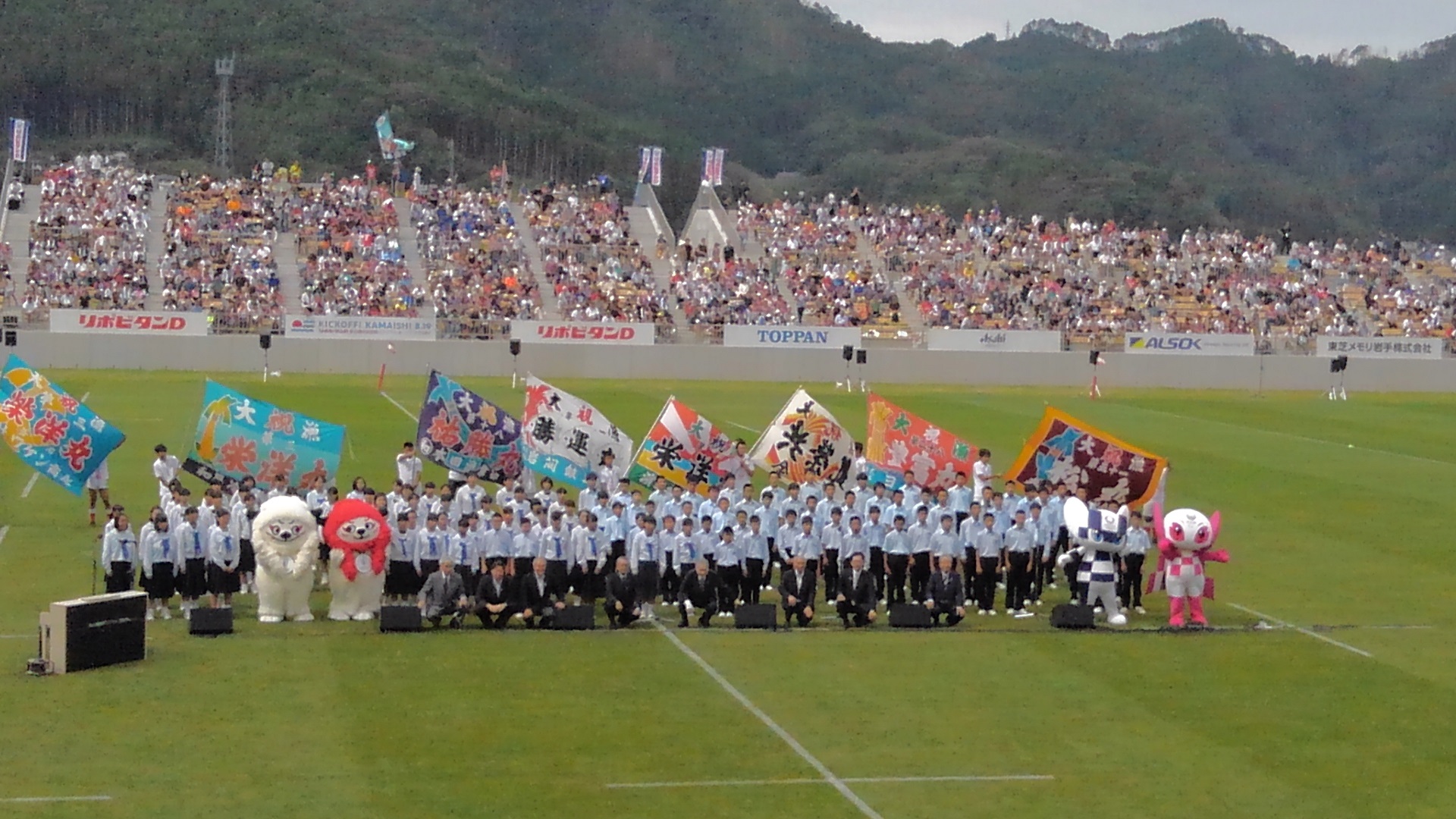 釜石鵜住居復興スタジアムの芝生のグラウンドの中心で、制服を着た学生たちが整列し、スーツを着た関係者たちがしゃがんでいる集合写真
