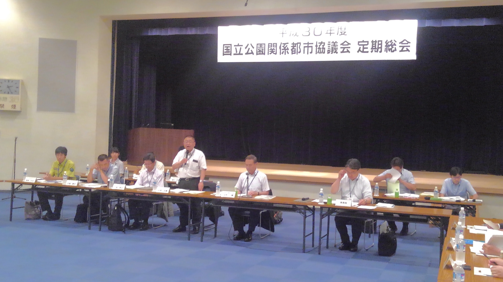 「国立公園関係都市協議会 定期総会」と書かれた横断幕が吊るされた舞台の前に置かれた長机の席の中央で、右手にマイクを持ち立ちながら話をする市長の写真