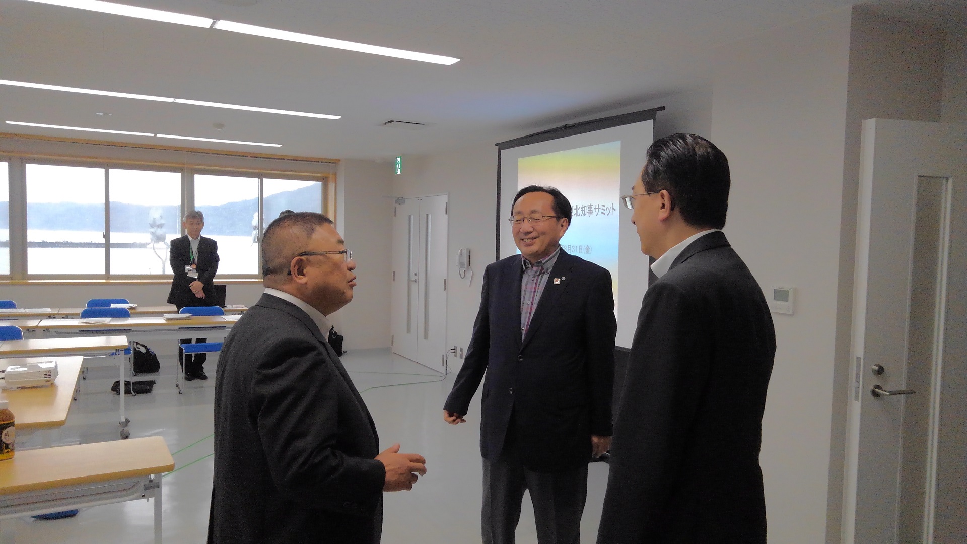 スクリーンが設置された部屋の中で、市長と2名の男性が立って話をしている写真