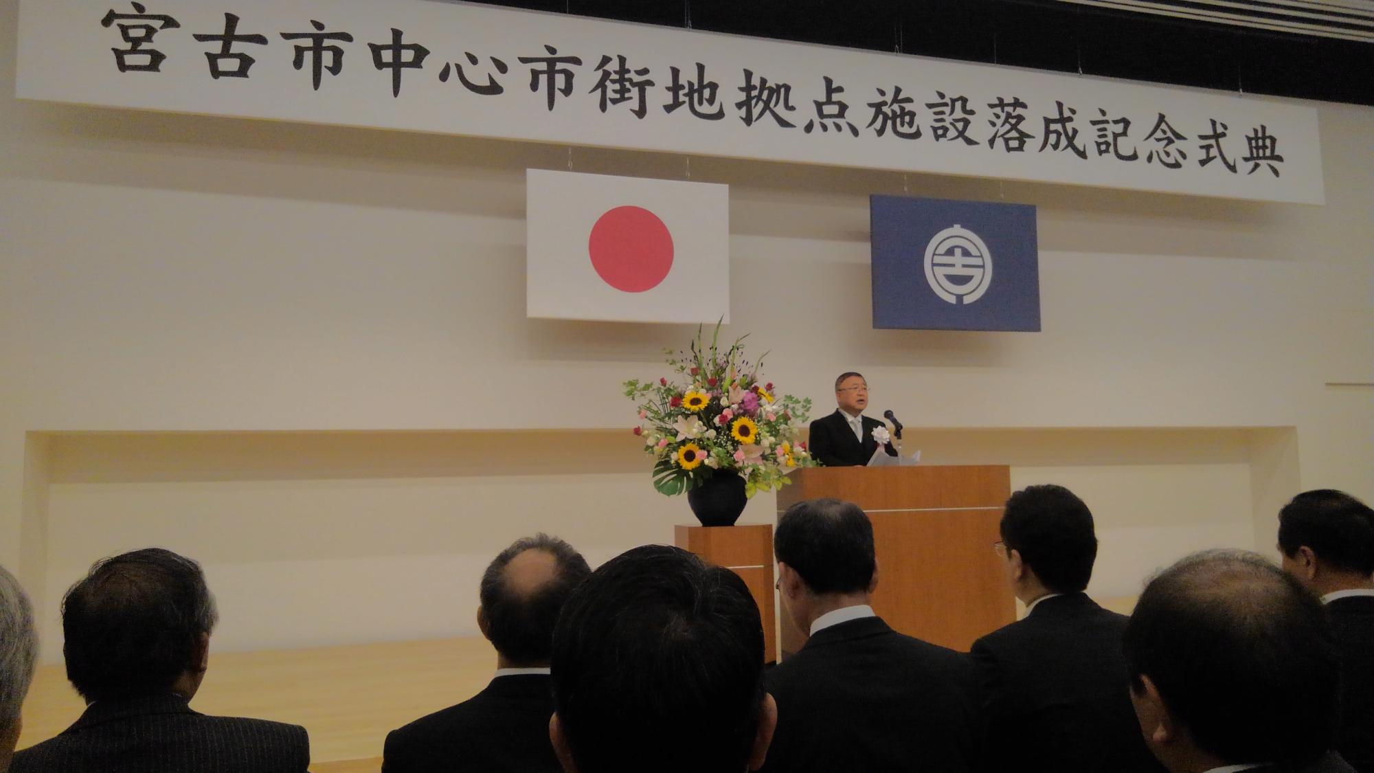 「宮古市中心市街地拠点施設落成記念式典」と書かれた横断幕と日本国旗、宮古市の市町村旗が掲げられた舞台上で、演台に置かれたマイクに向かい話をする様子の市長の写真