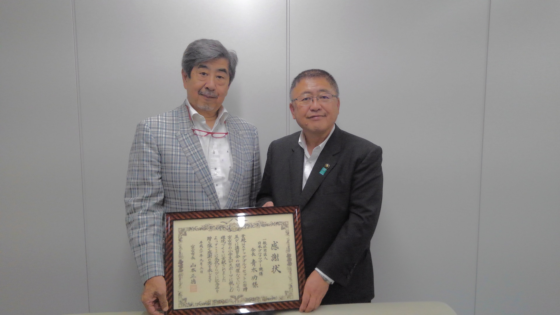 感謝状の額縁を一緒に持つ市長とグレーのチェック柄のスーツを着た男性の記念写真