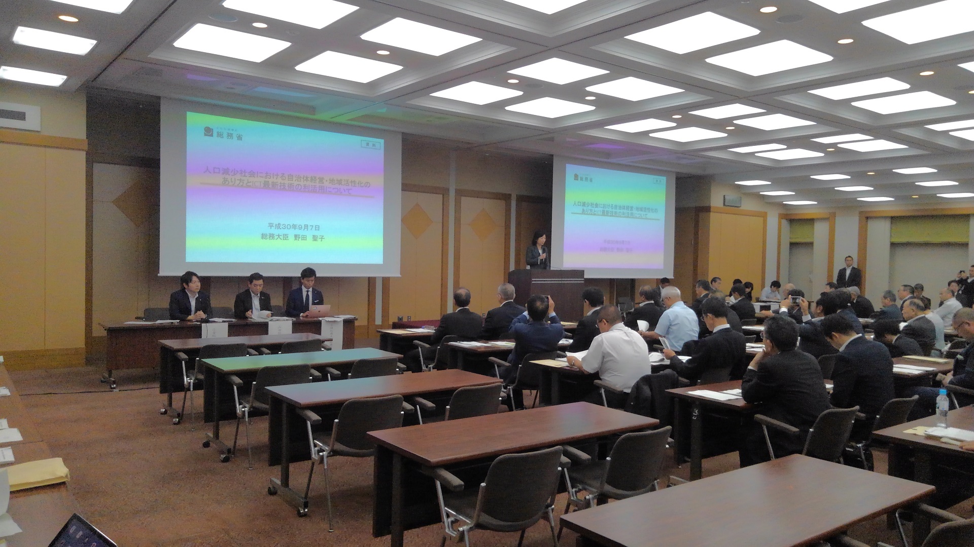 画面が映されたスクリーンが2台設置された会場で、並べられたテーブルに着き講演を聞く様子の参加者たちを斜め後方から写した写真