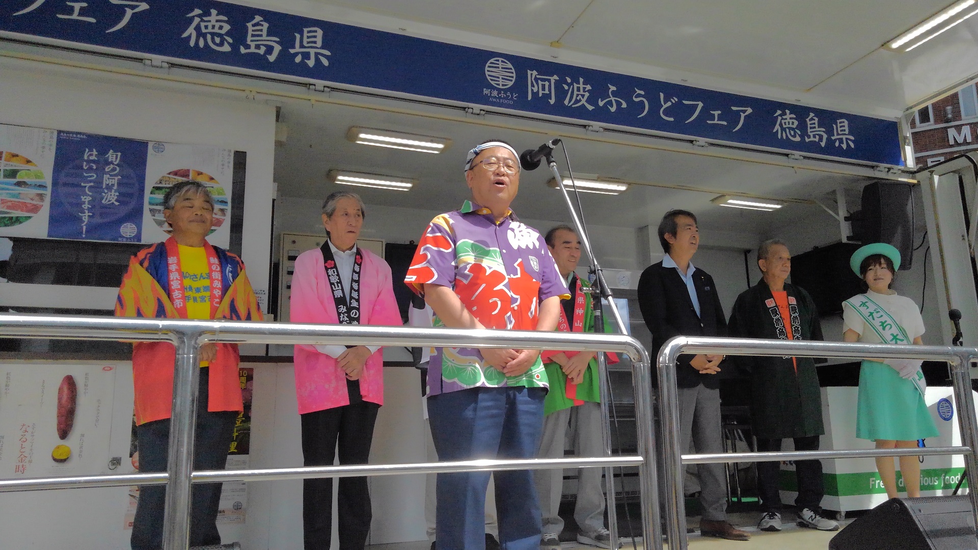スタンドマイクに向かって話をする様子の市長と、後方で立って並ぶ法被を着た方たちの写真