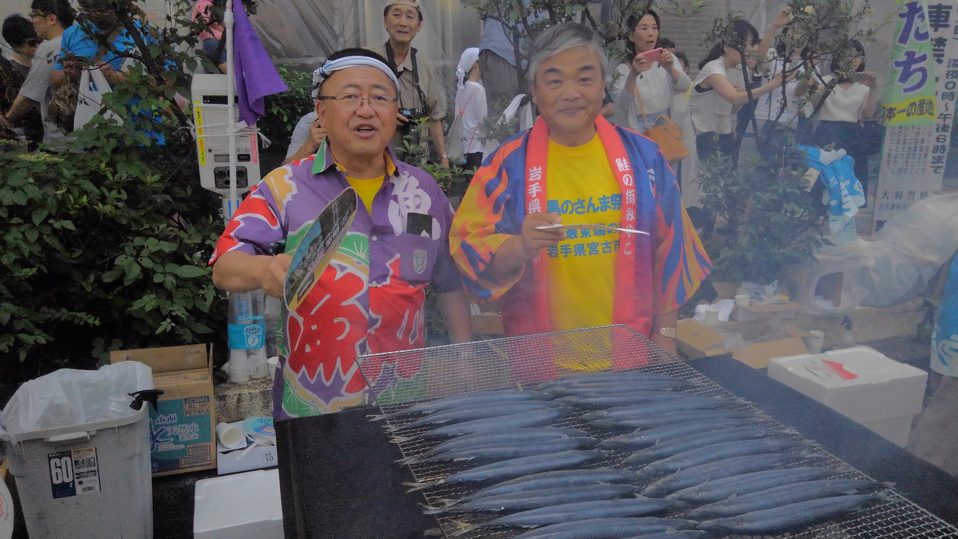 網の上に並べられ、煙を上げながら焼かれているサンマを団扇で仰ぐ市長と男性の写真