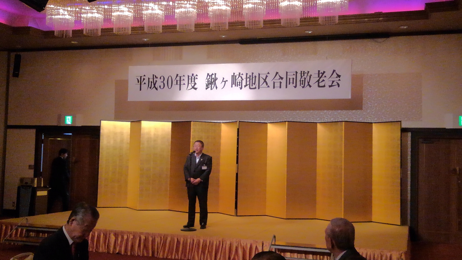 「平成30年度 鍬ヶ崎地区合同敬老会」と書かれた横断幕が吊るされ、金屏風が置かれた舞台上で、スタンドマイクに向かい話をする様子の市長の写真
