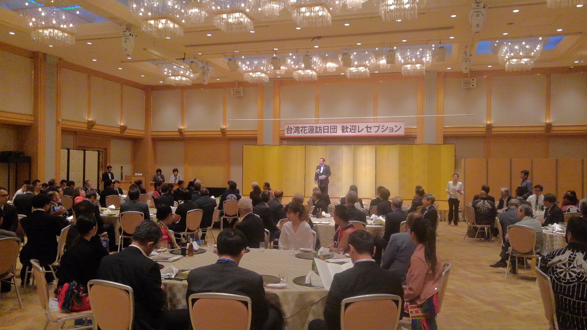 円卓が並べられた会場で、席に着く関係者の方々と金屏風の前で話をする様子の男性の写真