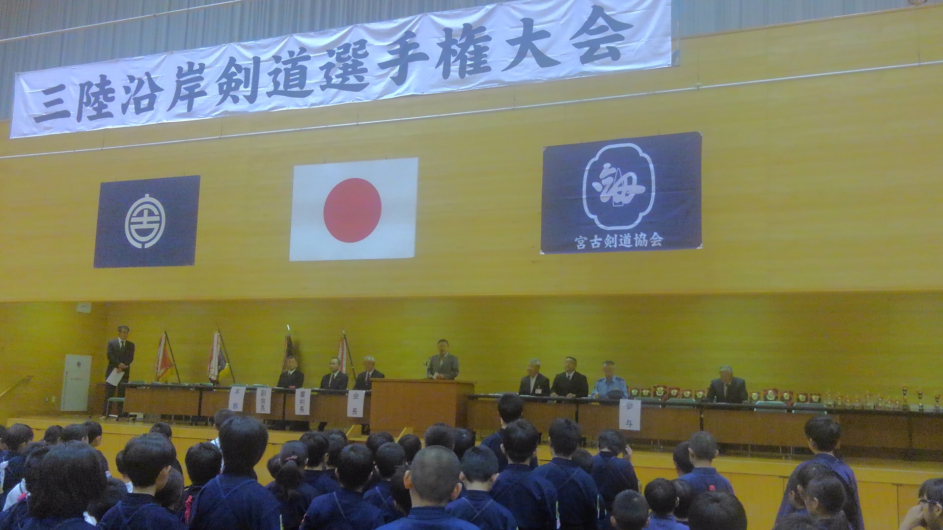 「三陸沿岸剣道選手権大会」と書かれた横断幕と宮古市の市町村旗、日本国旗、宮古剣道協会の旗が吊るされた会場で、整列する選手たちの前方に置かれた演台で話をする様子の市長の写真