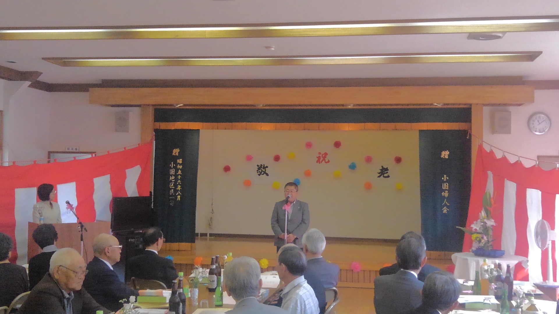 舞台後方の壁に装飾が施され、紅白幕が左右に垂らされた会場で、スタンドマイクに向かい話をする様子の市長