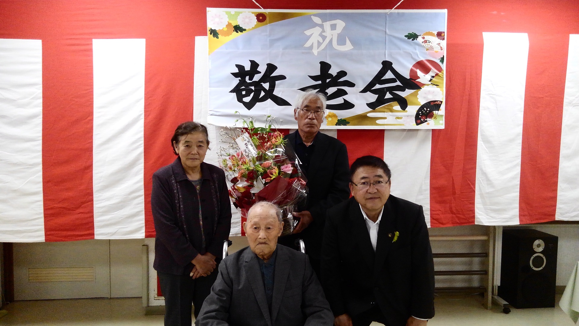 「祝 敬老会」と書かれた横断幕が吊るされた紅白幕の前で、椅子に座る市内最高齢者の川端浅吉さんとその隣で中腰で立つ市長、後方で立つ女性と花束を持った男性の記念写真