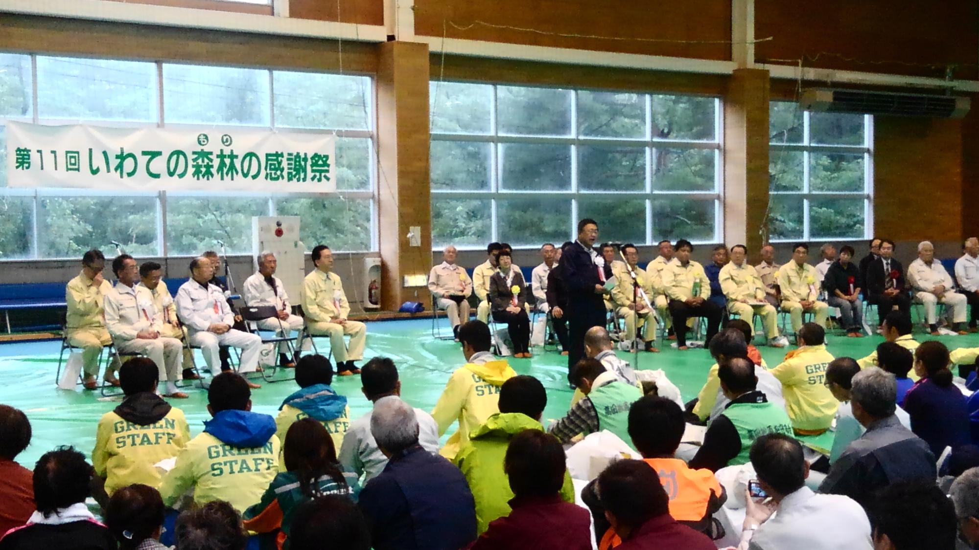 「第11回いわて森林の感謝祭」と書かれた横断幕が窓際に吊るされた会場で、作業服を着て椅子に座る関係者の方々の前で、スタンドマイクに向かい話をする様子の市長と、向かい側で座って話を聞く参加者たちの写真