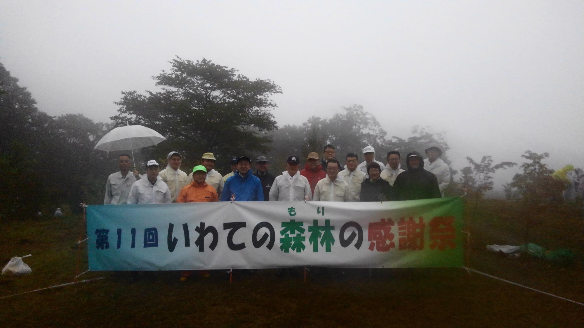 霧が立ち込める森の中で、「第11回いわて森林の感謝祭」という横断幕を持って2列に並んで記念撮影をする市長や参加者たちの写真