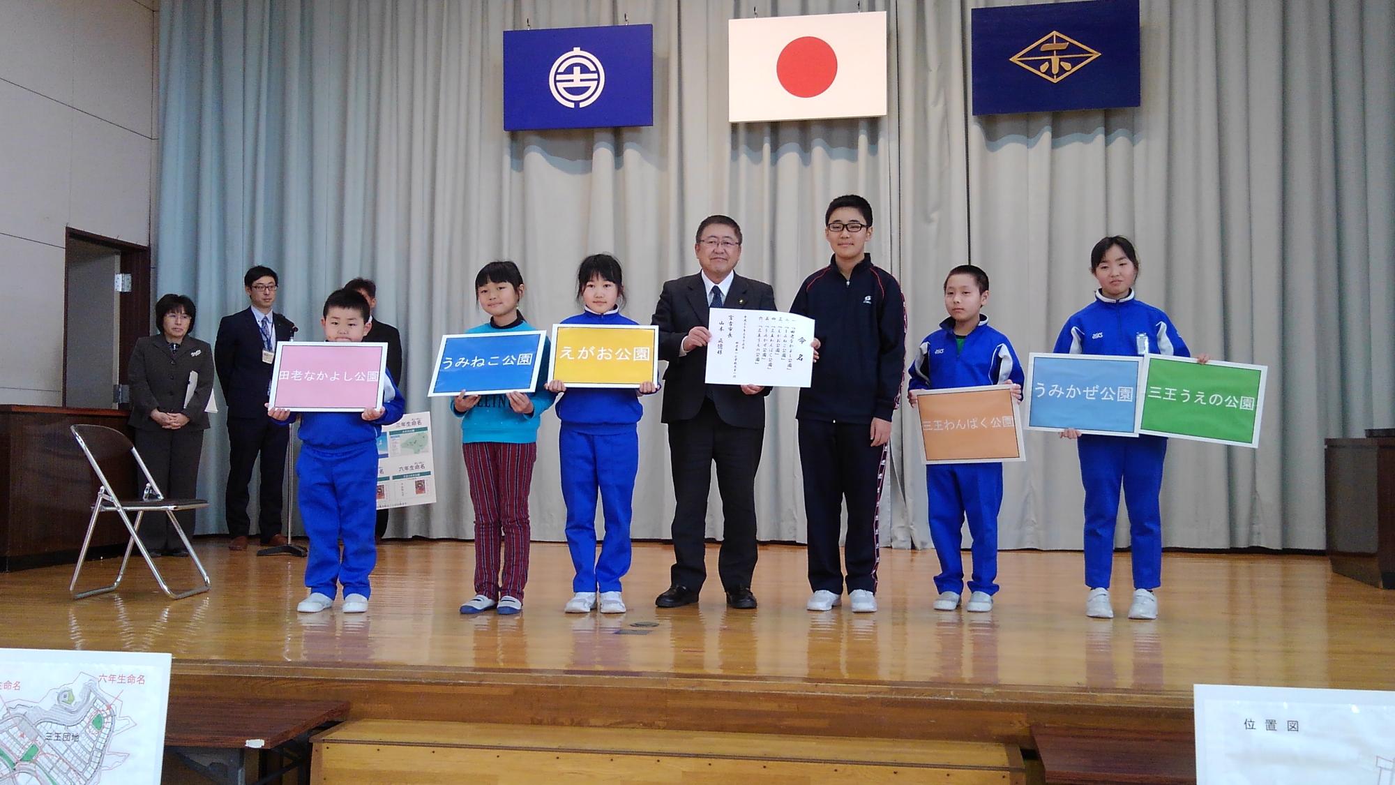 ジャージを着た小学生6名と市長が、パネルを持って舞台上に横一列に並んでいる写真