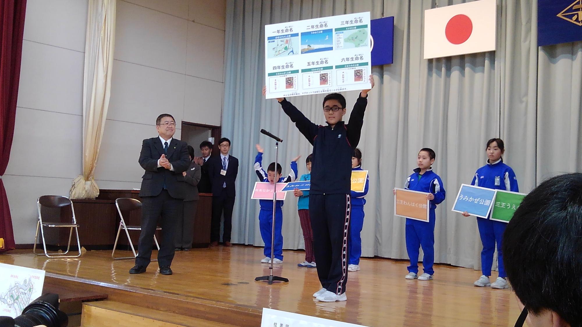 舞台上で絵や文字が書かれたパネルを両手で持ち上に掲げる男の子の写真