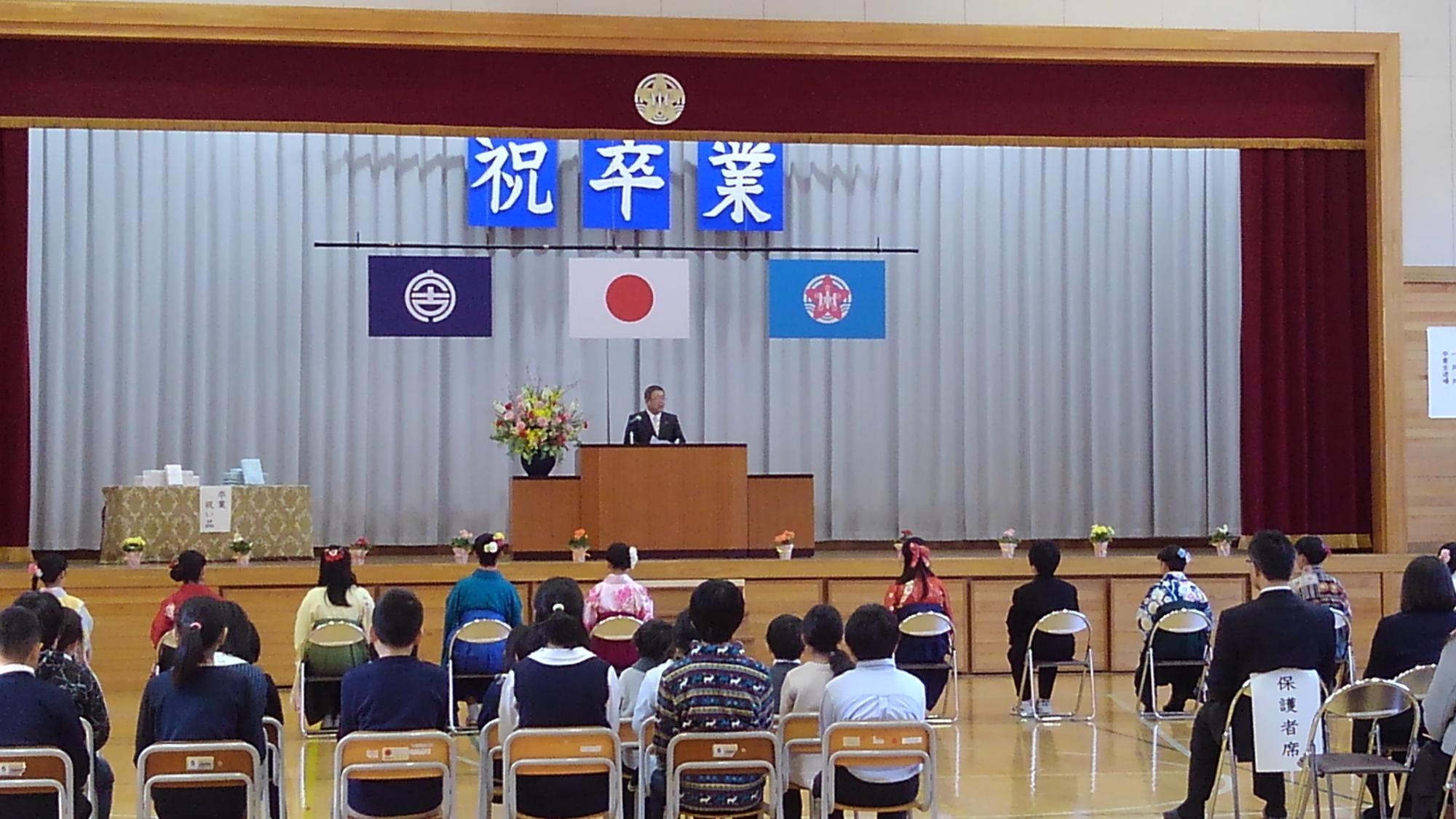 「祝卒業」と書かれたパネルが吊るされた舞台上の演台の前に立つ市長と、袴を着てパイプ椅子に座り横一列に並んだ卒業生、後方で椅子に座る在学生と保護者たちの写真