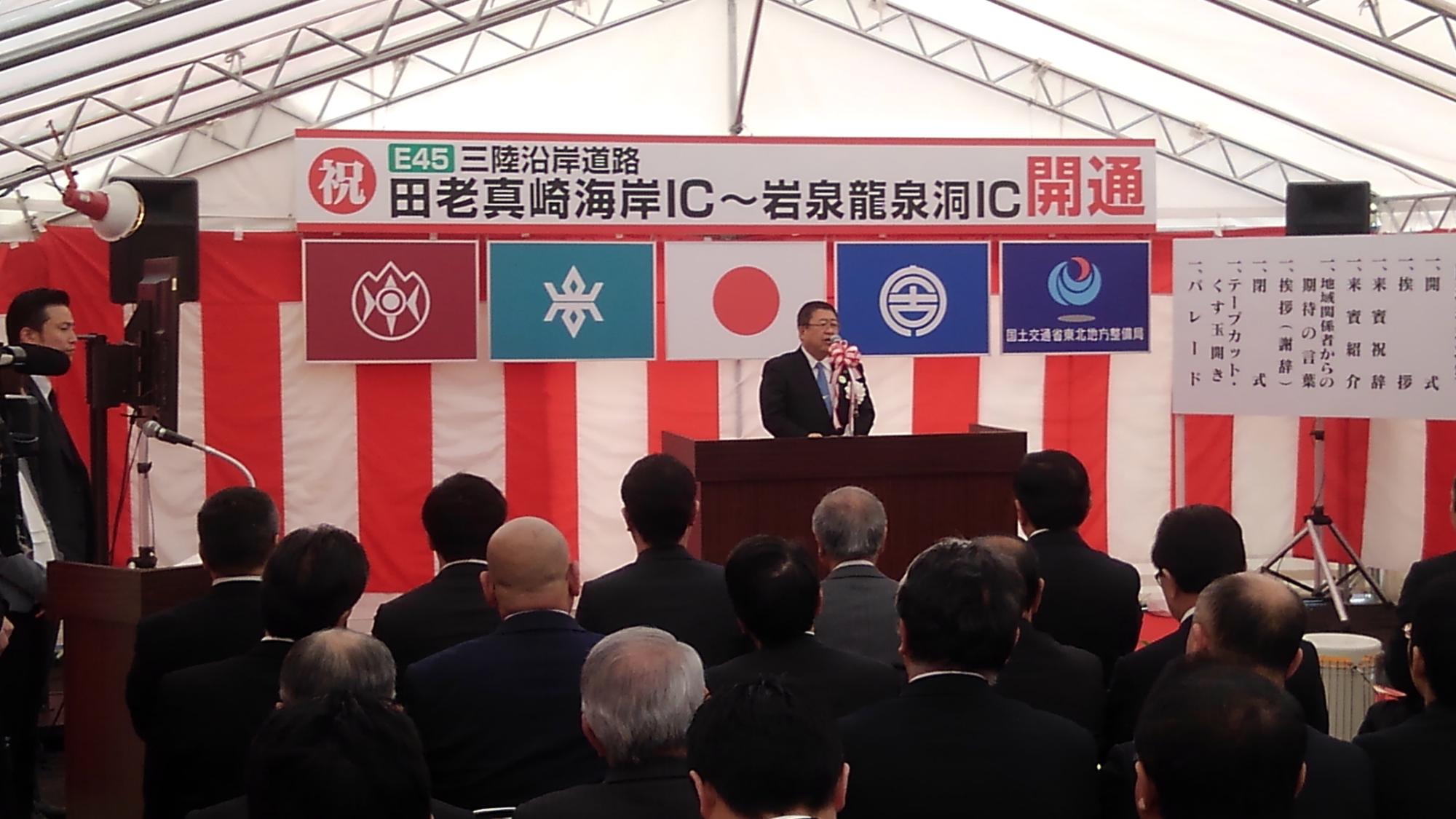 紅白幕が貼られた壁の前で、演台に置かれたマイクに向かい話をする様子の市長の写真