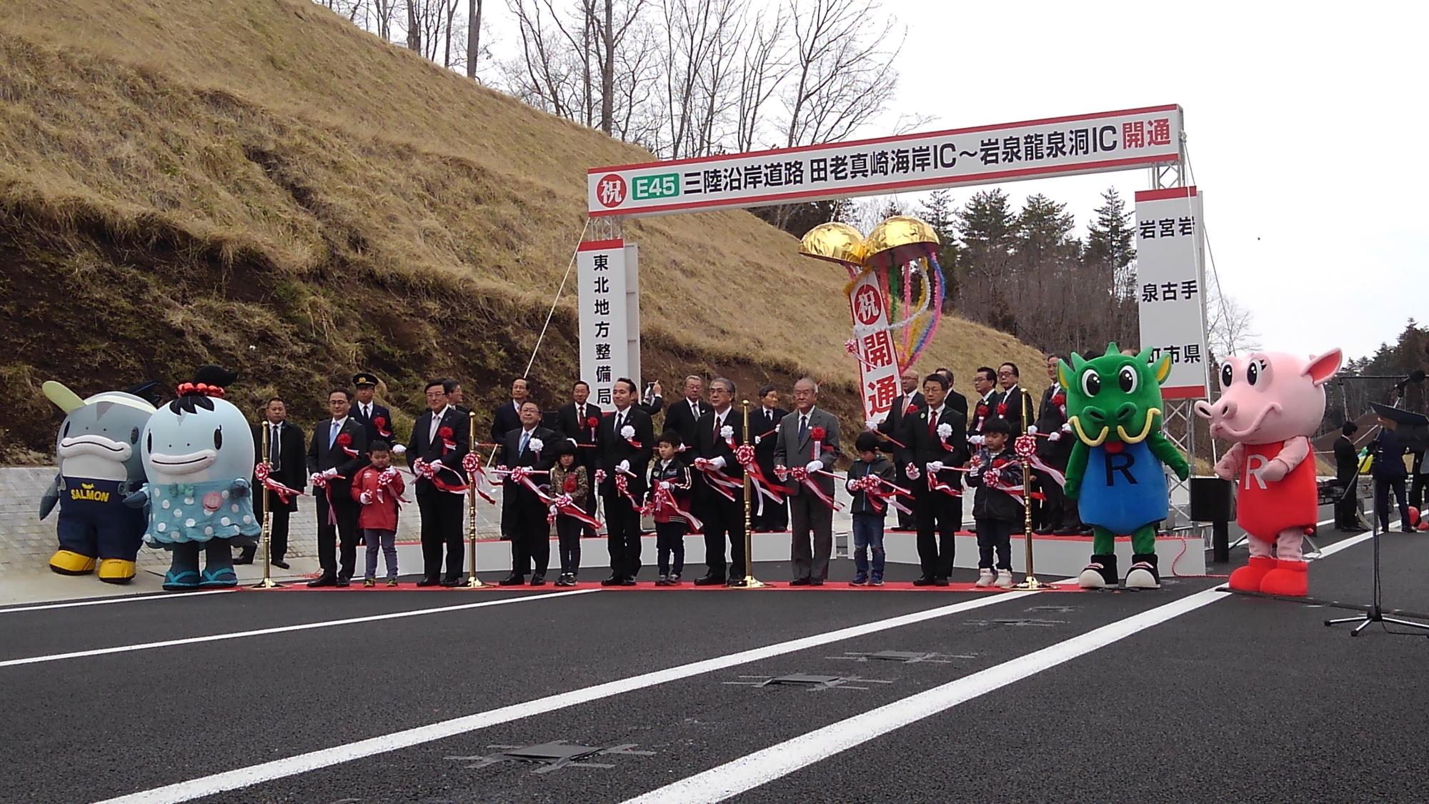 道路を横断するように敷かれたレッドカーペットの上で、テープカットをする関係者の男性と子供たち、サーモンくんとみやこちゃん、龍ちゃんと泉ちゃんの写真