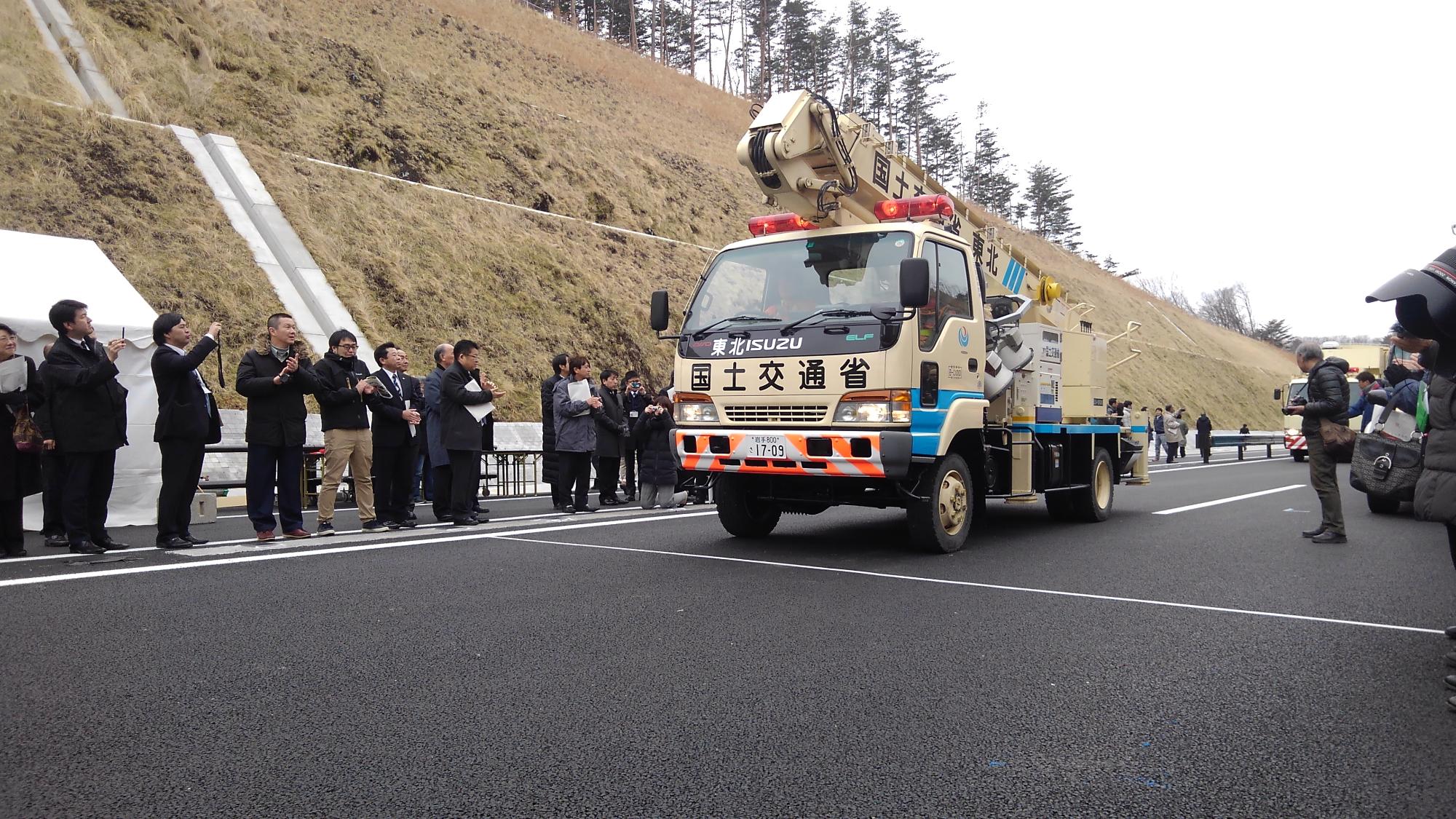 「国土交通省」と書かれたトラックが道路を進む様子を道路わきから見ている関係者の方々の写真