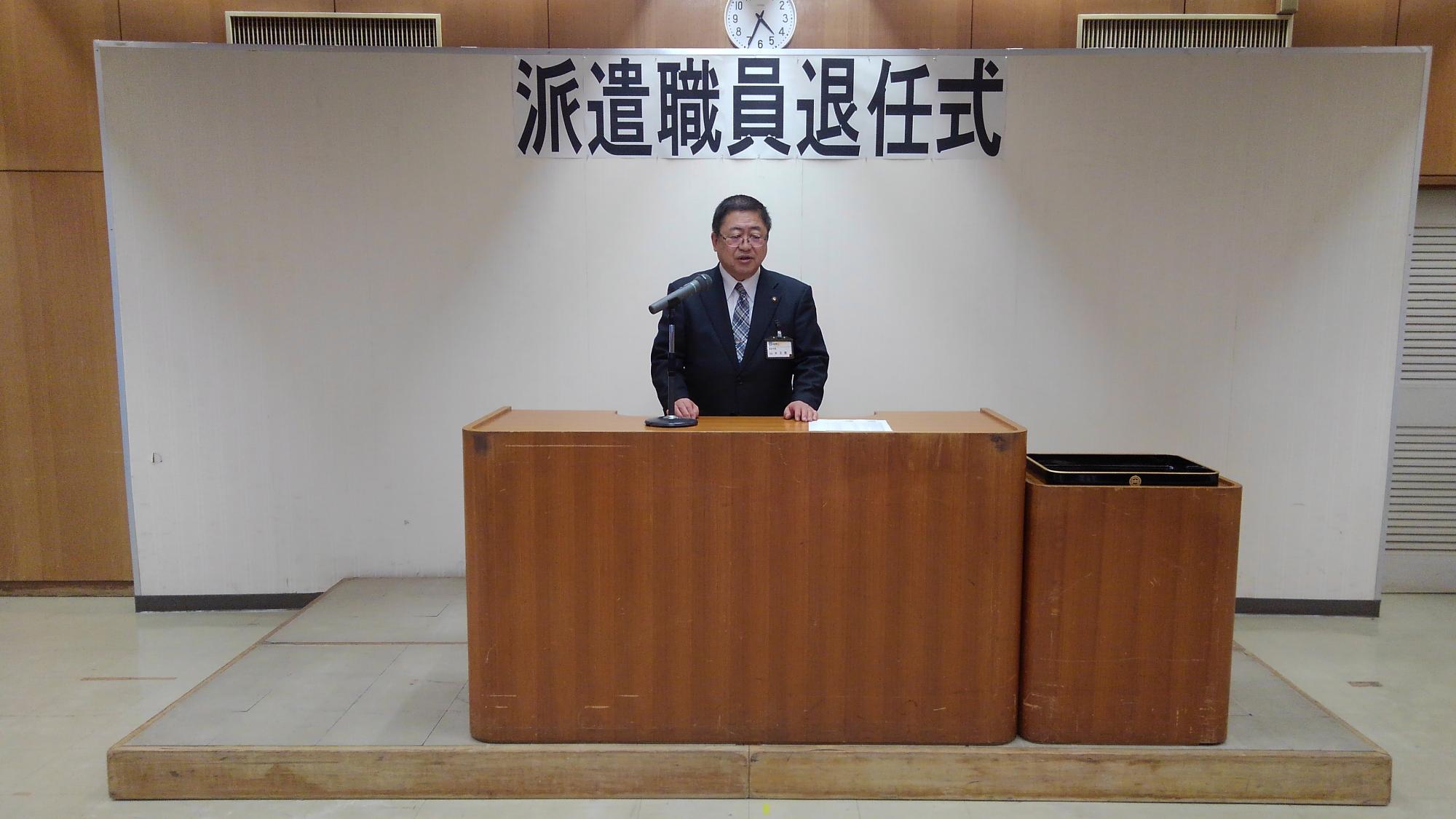 「派遣職員退任式」と書かれた紙が貼られたボードの前で、演台に置かれたマイクに向かい話をする様子の市長の写真