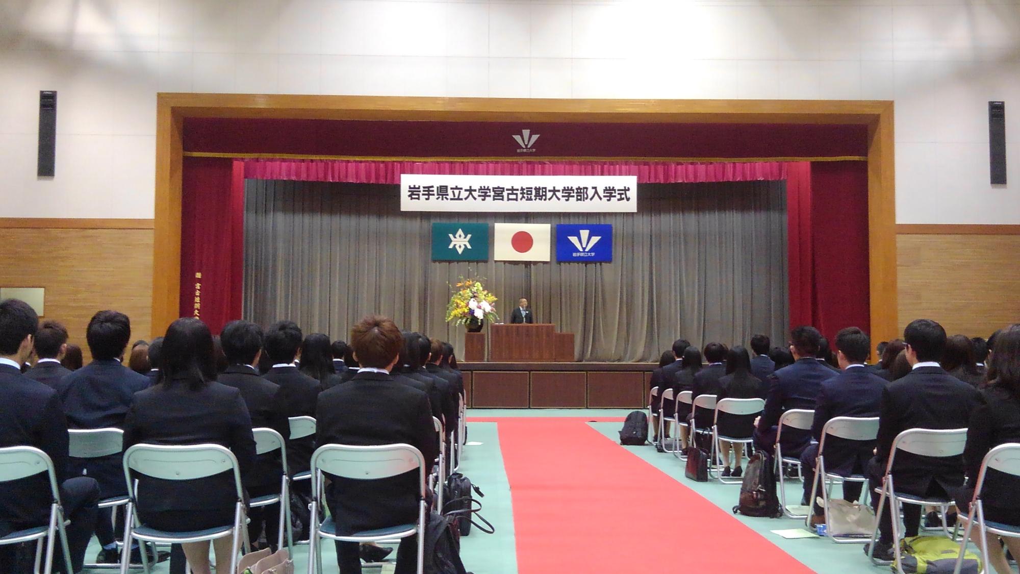 並べられた椅子に座り整列する入学正たちと、舞台上に置かれた演台で話をする様子の市長の写真