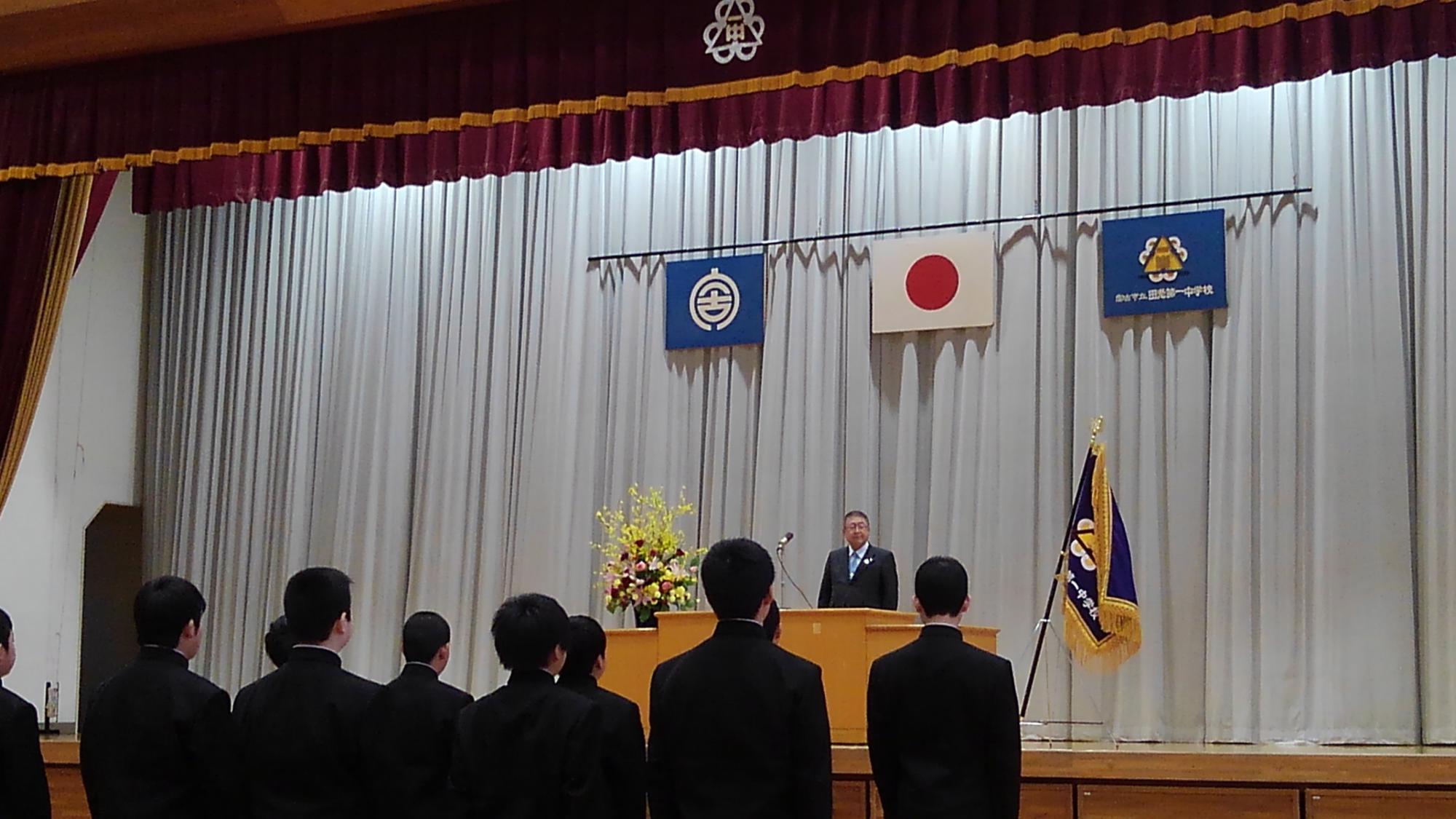 立って整列する制服を着た新入生の前で、舞台上に置かれた演台に向かい話をする様子の市長の写真