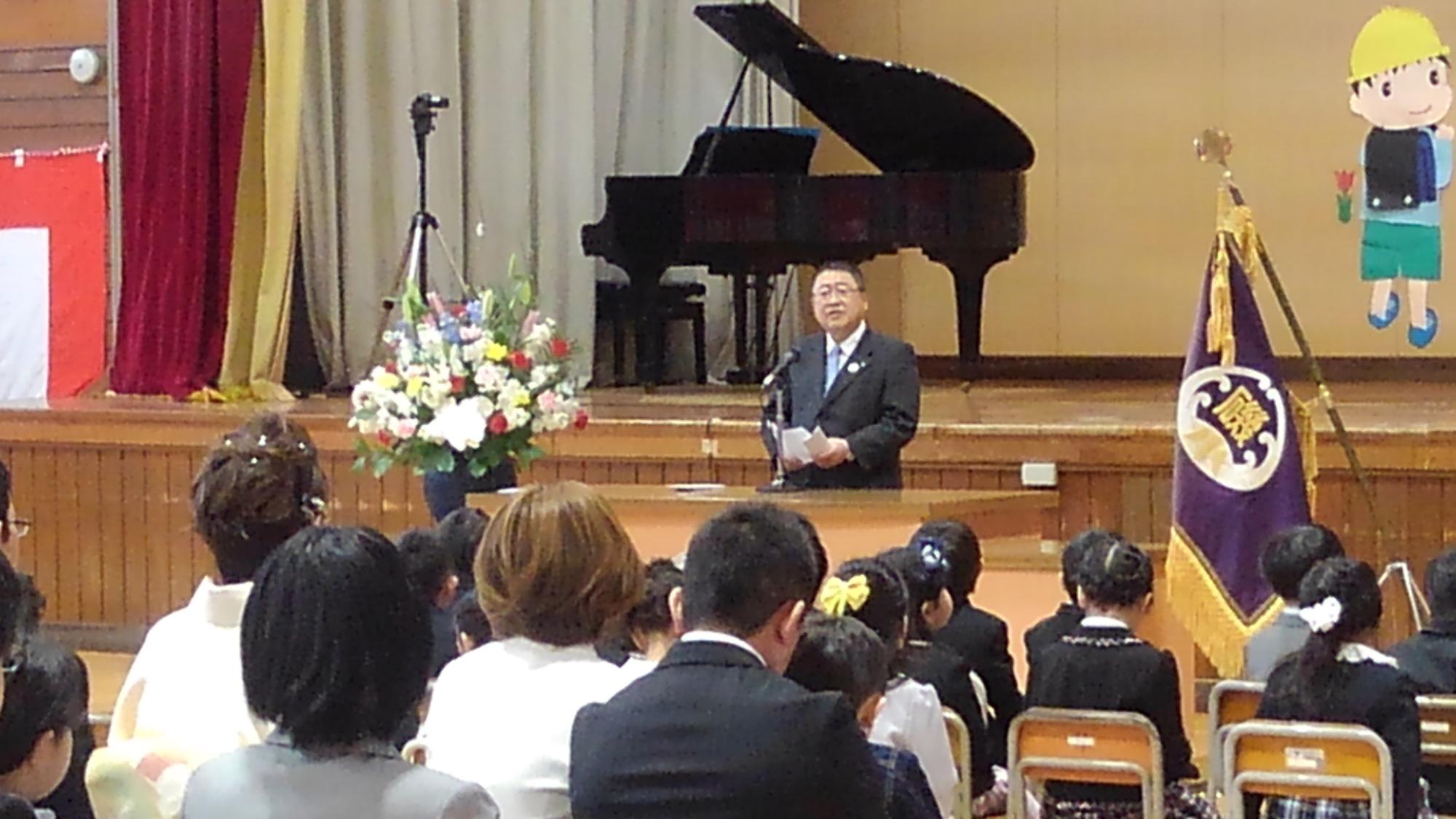 椅子に座った新入生や保護者の前で演台のに置かれたマイクに向かい話をする市長の写真