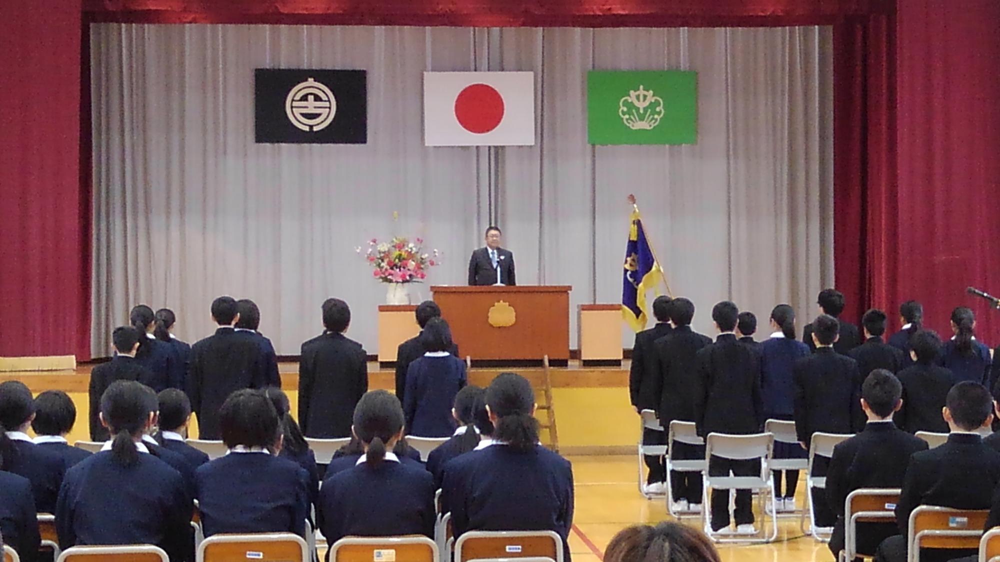 舞台上に置かれたマイクに向かい話をする市長と、並べられた椅子の前に立つ制服を着た新入生たちの写真
