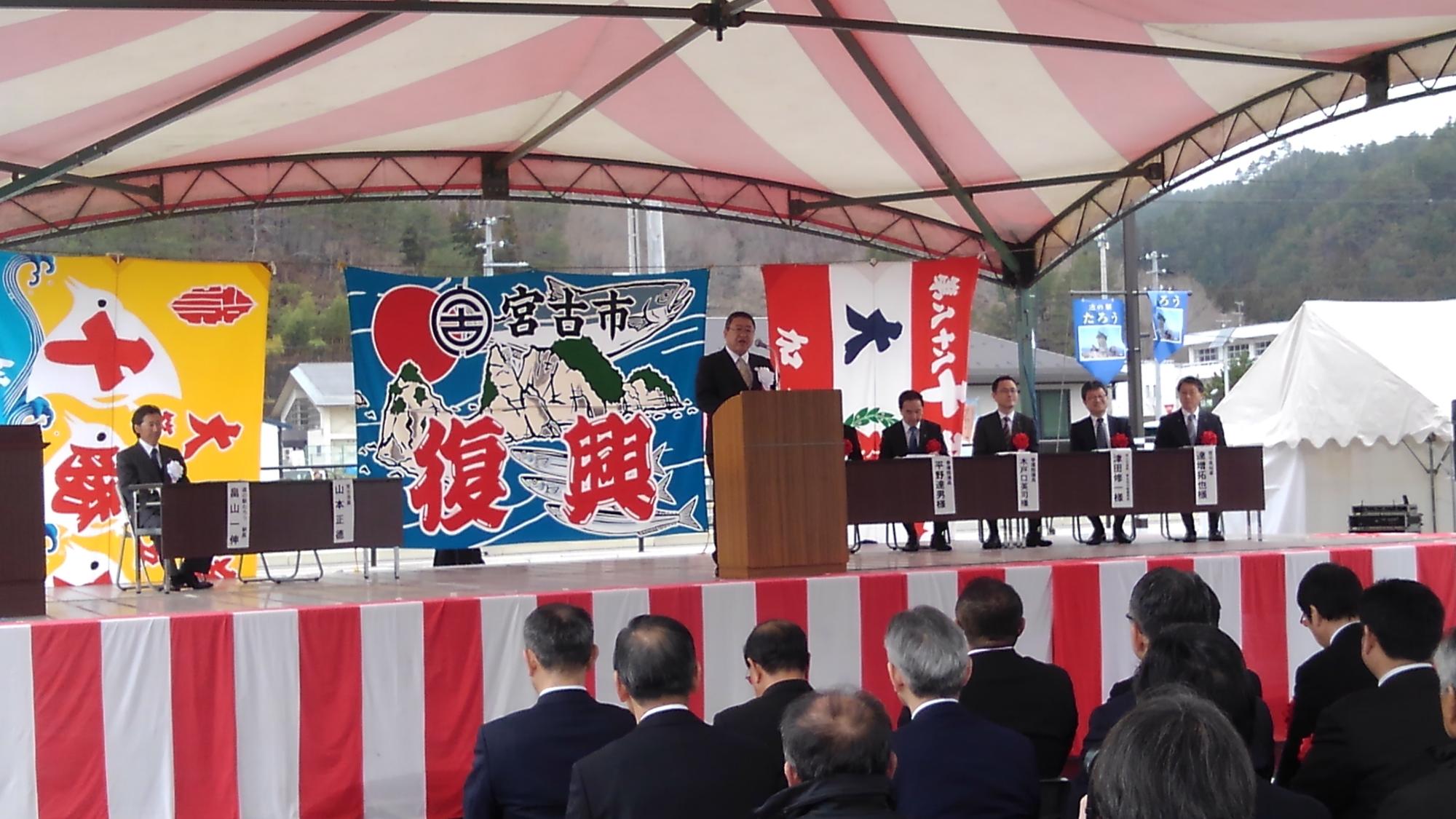 大漁旗が掛けられた舞台で、演台に置かれたマイクに向かい話をする様子の市長の写真