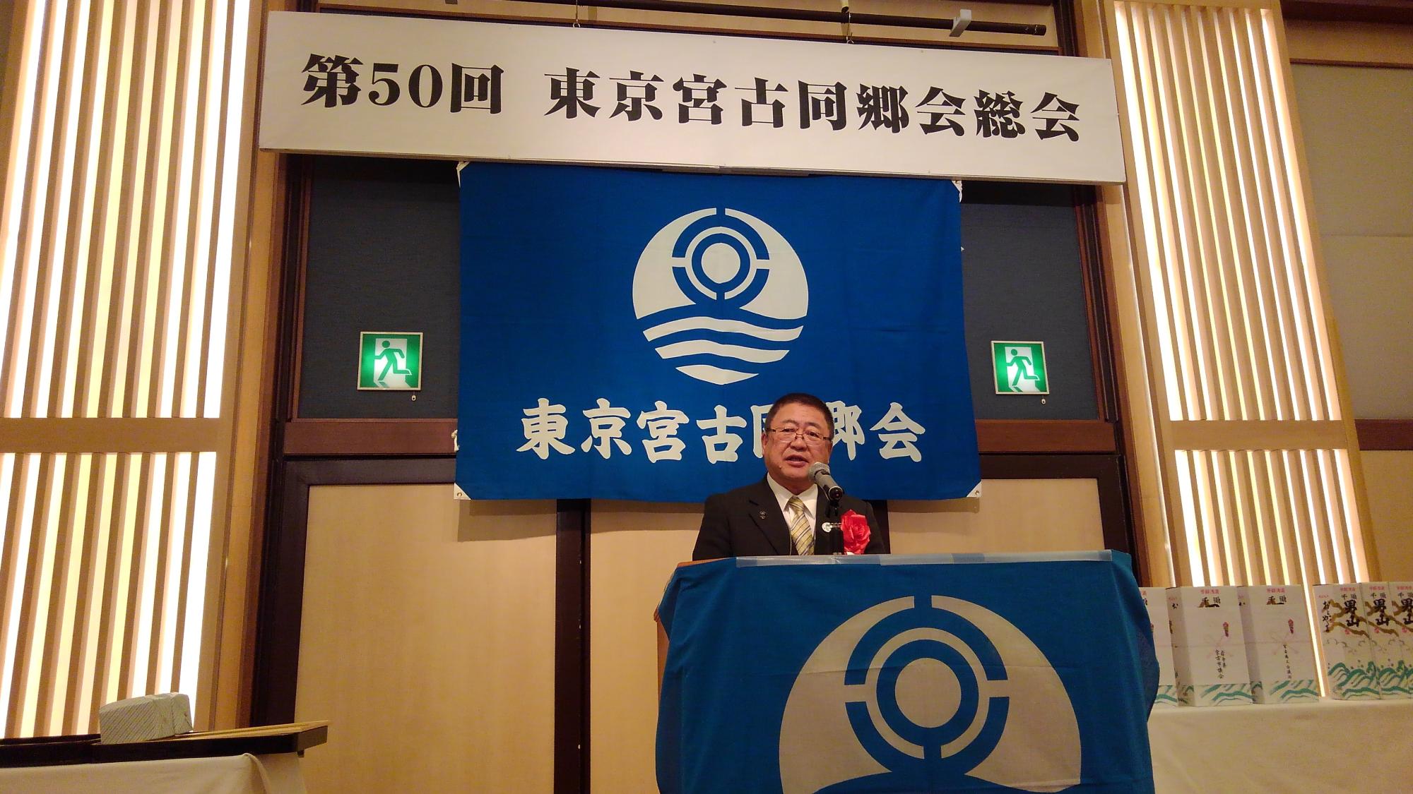「第50回 東京都同郷会総会」と書かれた横断幕が吊るされた会場で、演台に置かれたマイクに向かい話をする様子の市長の写真