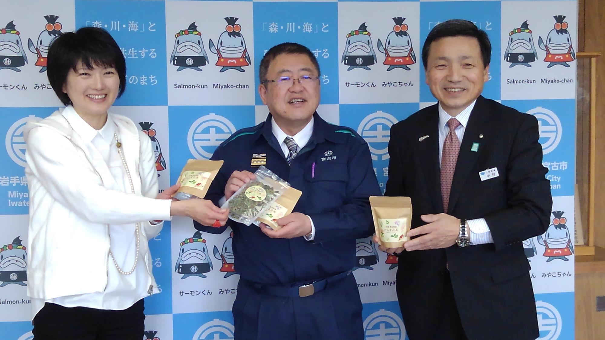 バックパネルの前でたらま花茶の袋を持つ市長と女性1名と男性1名の記念写真