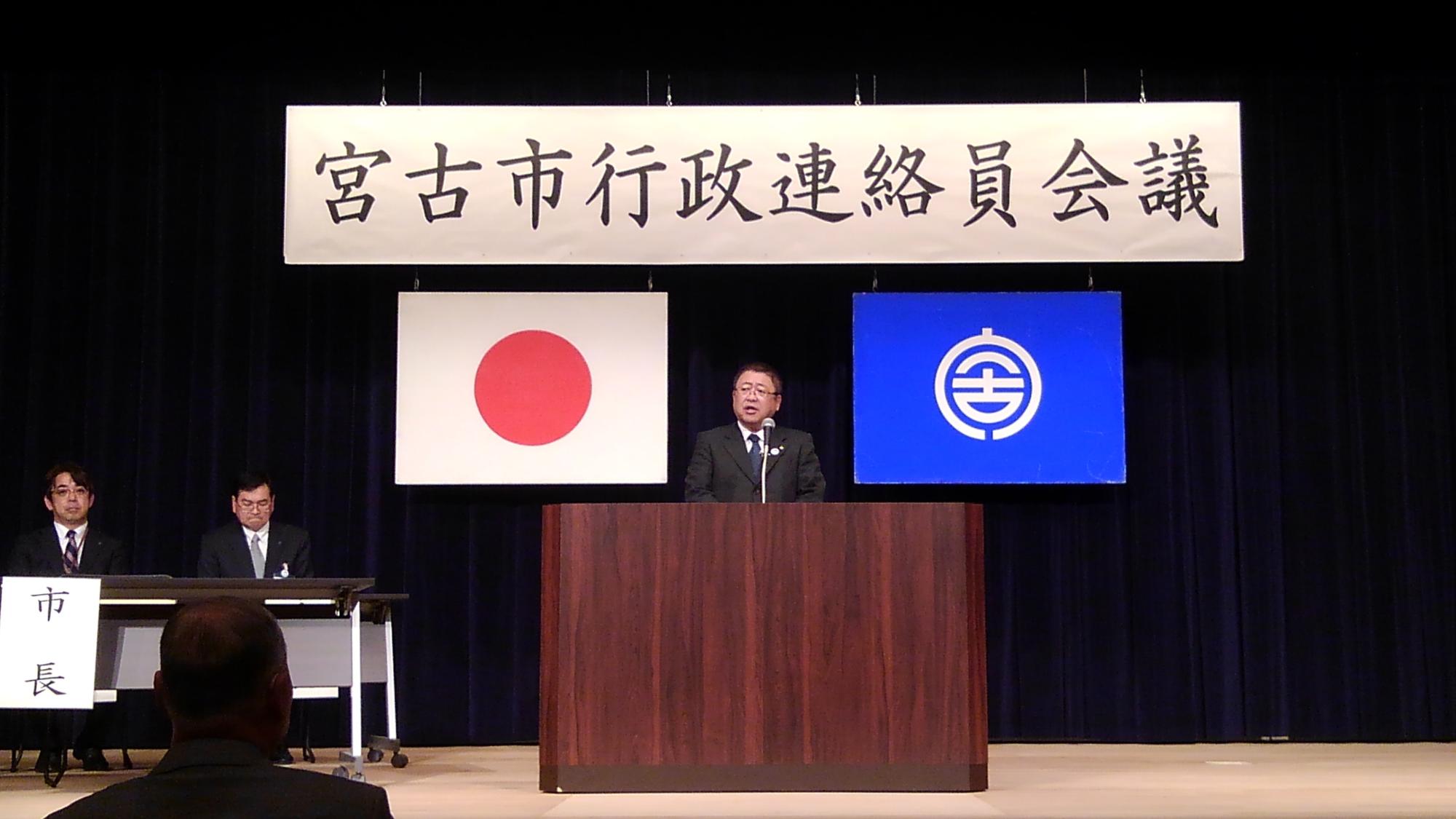「宮古市行政連絡員会議」と書かれた横断幕、日本国旗、宮古市の市町村旗が吊るされた舞台上の演台に置かれたマイクに向かい話をする市長の写真