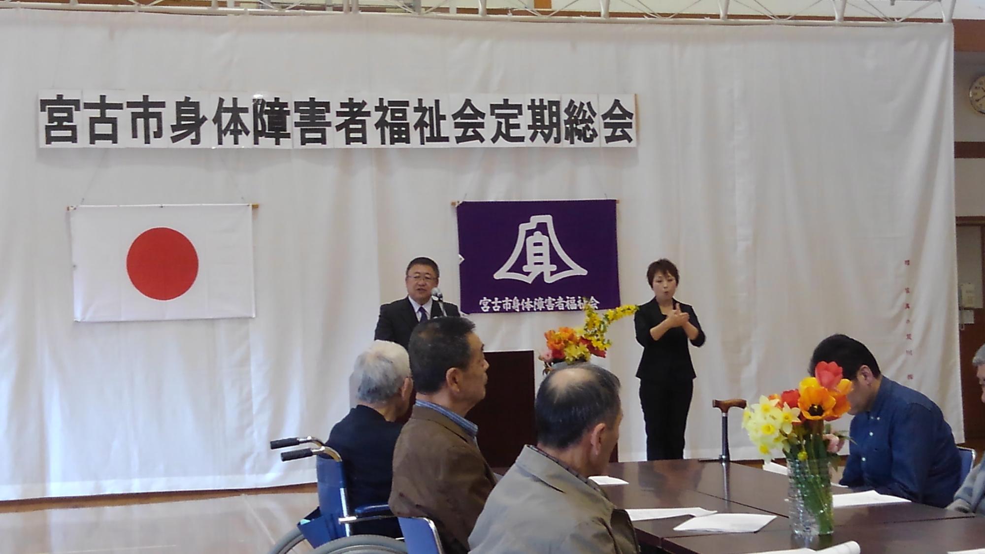 「宮古市身体障害者福祉会定期総会」と書かれた紙が掛けられた壁の前で、演台に置かれたマイクに向かい話をする市長と、テーブルに向かい合うように座る参加者たちの写真