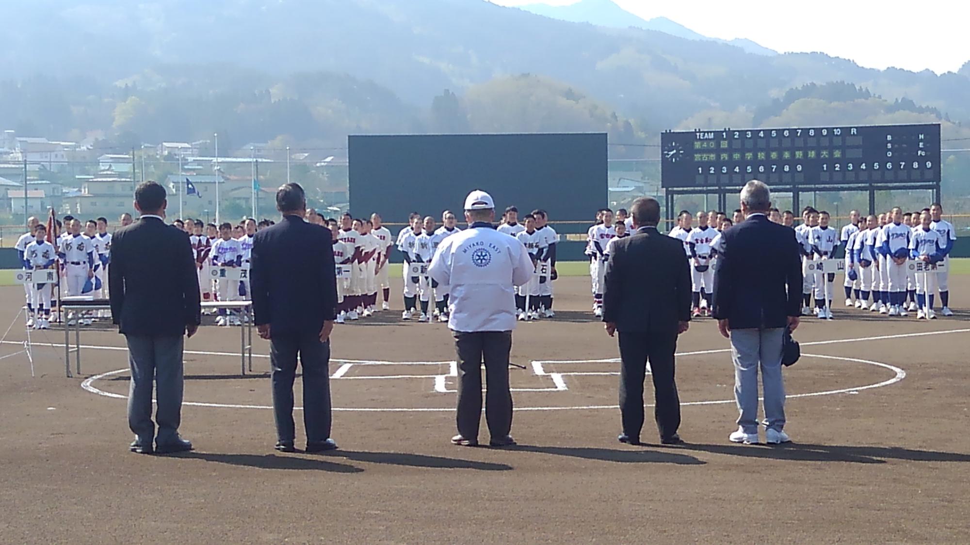 野球グラウンドでユニフォームを着て整列する選手たちの向かいで整列する5名の関係者の男性たちの写真