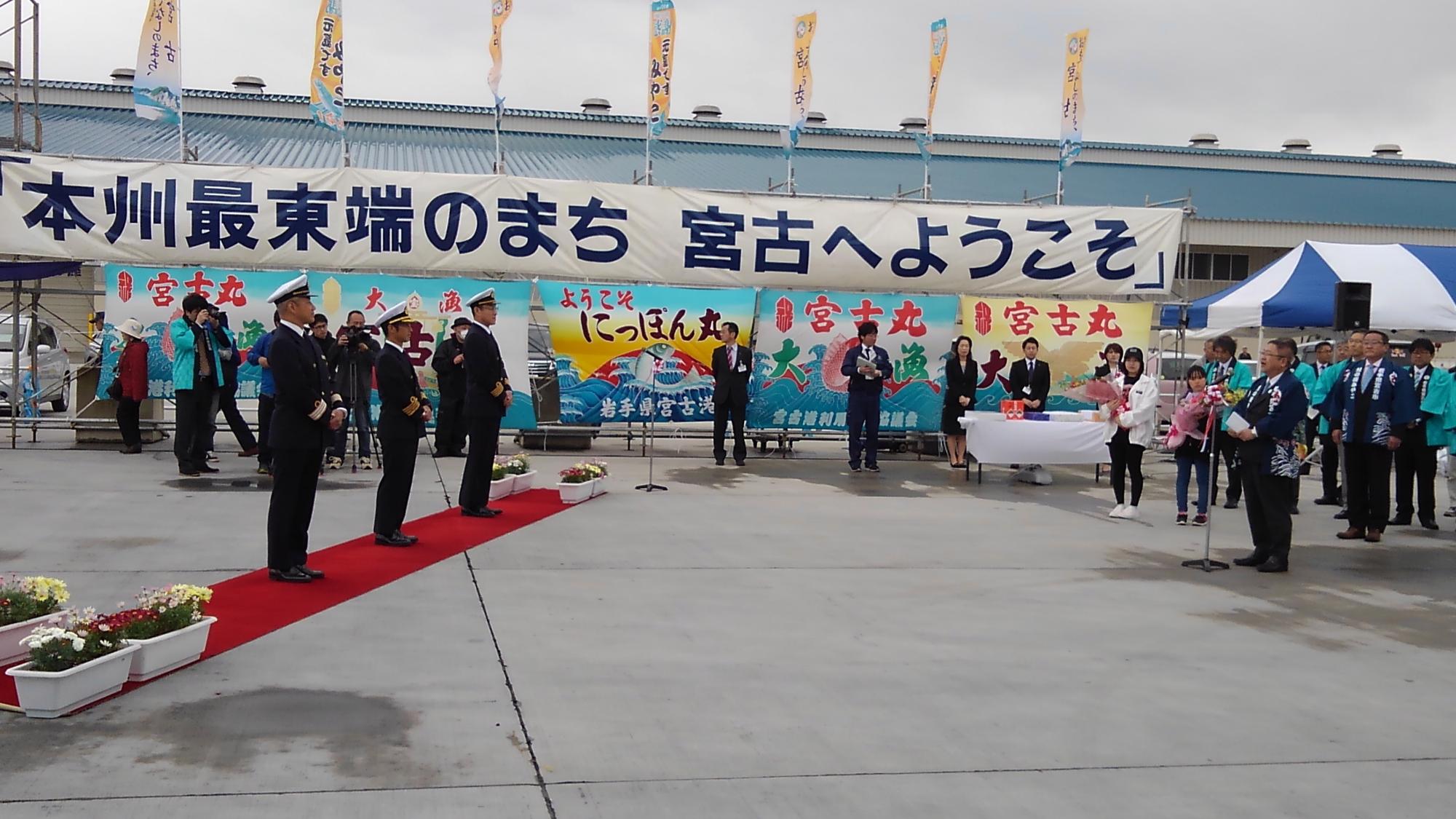 「本州最東端のまち 宮古へようこそ」と書かれた横断幕と大漁旗の前で、レッドカーペットの上に並ぶ3名のにっぽん丸の乗組員の方と、向かい側で法被を着てスタンドマイクに向かい話をする様子の市長の写真