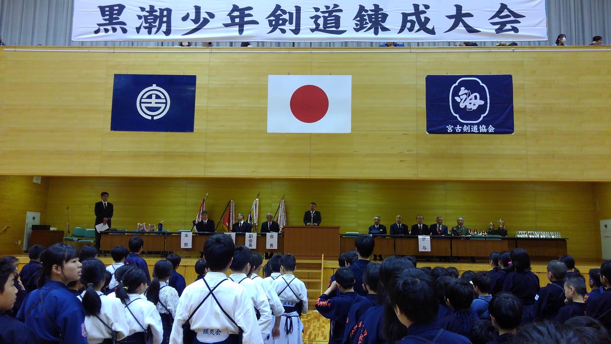 「黒潮少年剣道錬成大会」と書かれた横断幕、宮古市の市町村旗、日本国旗、宮古市剣道協会の旗が掲げられた会場で、剣道着を着て整列する選手たちと演台で話をする様子の市長の写真