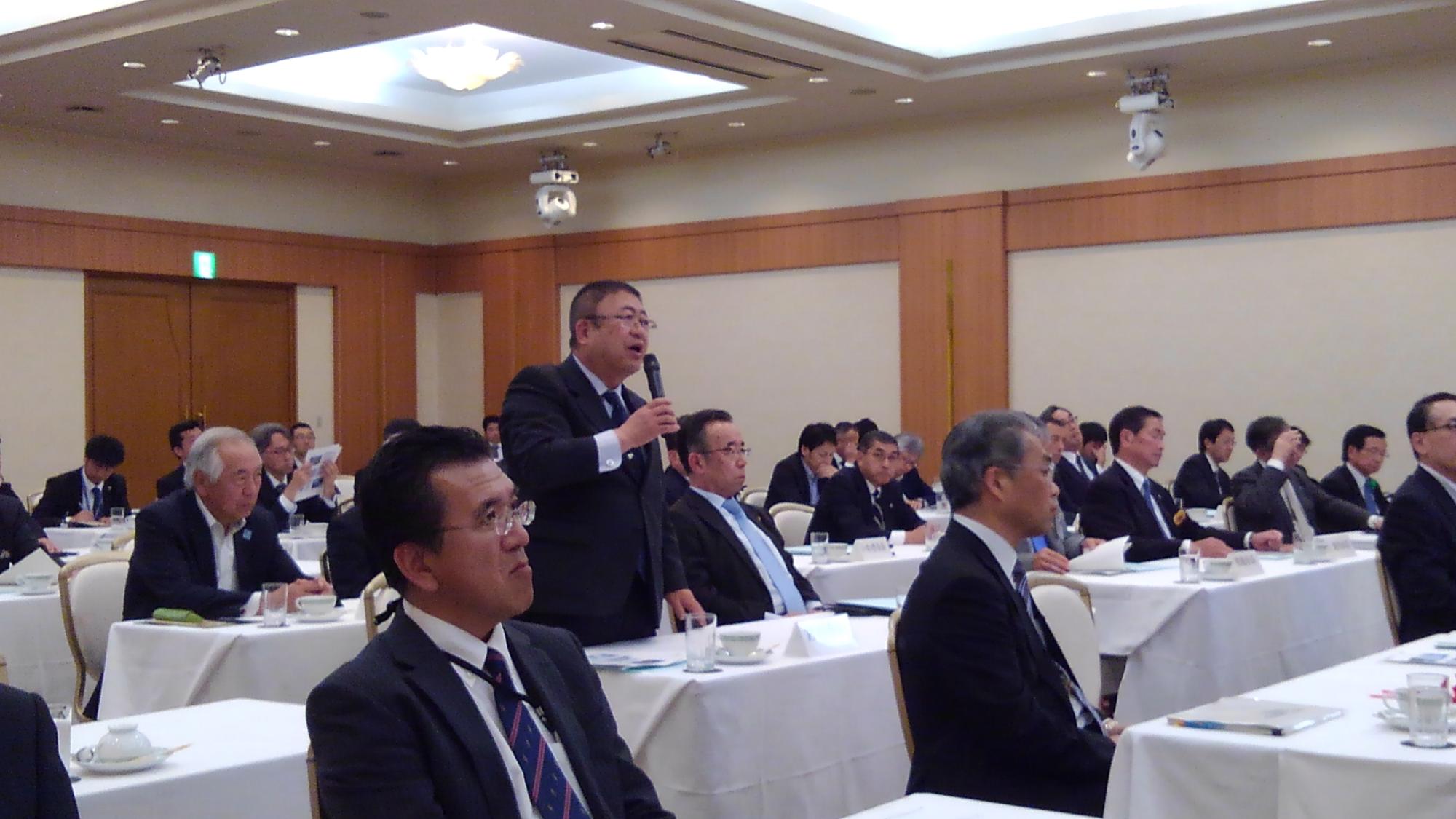 白いテーブルクロスが敷かれた机が並んだ会場で、着席する多くの関係者の中央で立って右手にマイクを持ち話をしている様子の市長の写真