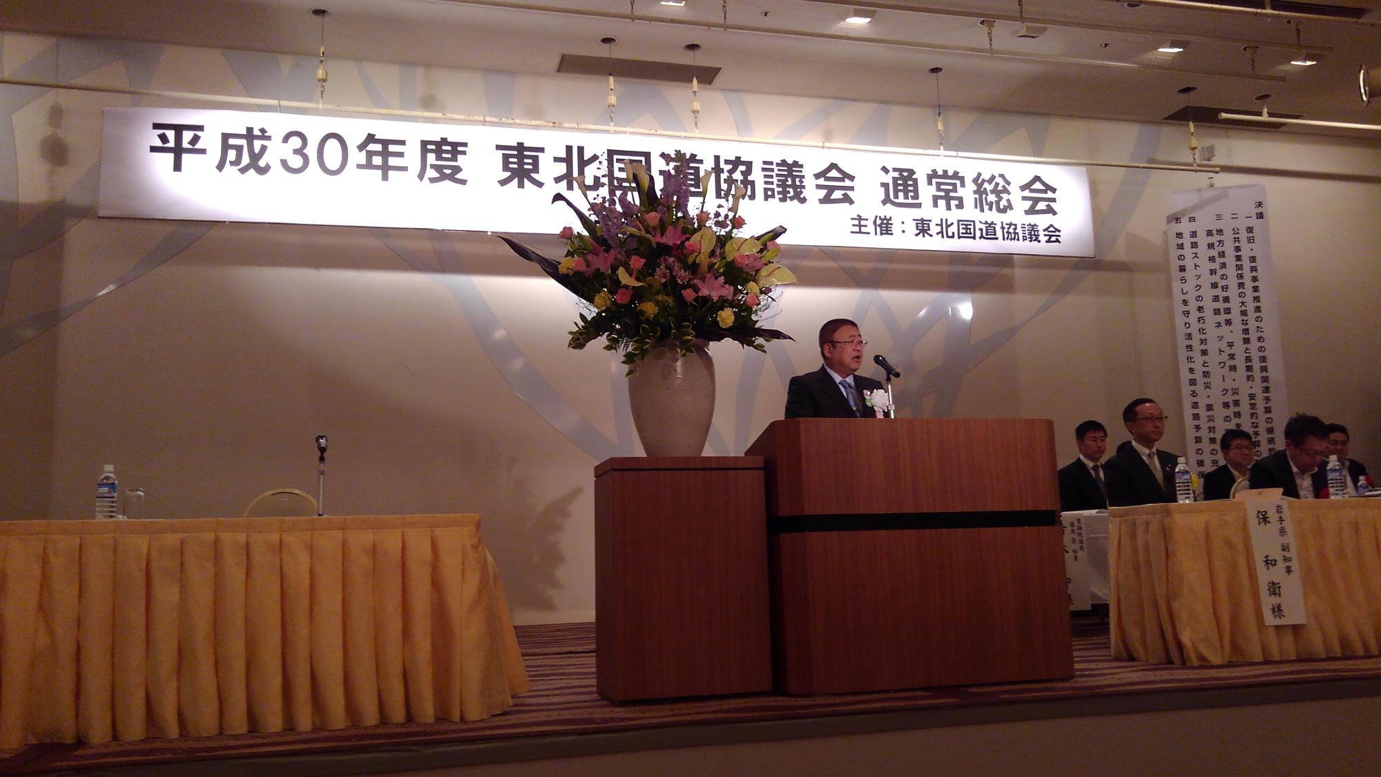 「平成30年度 東北国道協議会 通常総会」と書かれた横断幕が吊るされた舞台上で、たくさんの花が活けられた大きな花瓶が置かれた台の横の演台で話をする様子の市長の写真