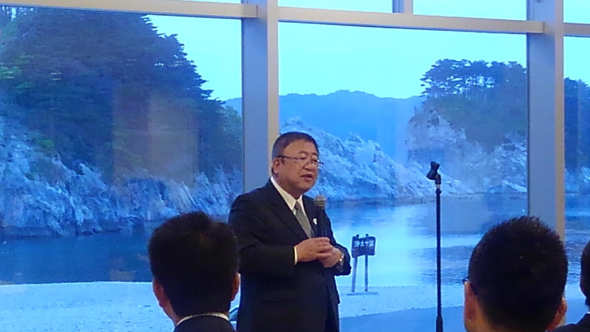 ガラス張りの建物の中で、両手でマイクを持ち話をする様子の市長の写真