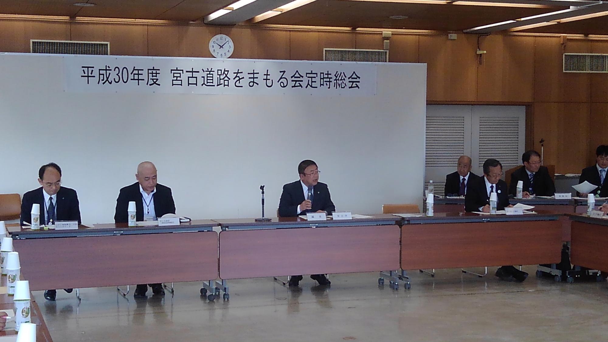 「平成30年度宮古道路をまもる会定時総会」と書かれた紙が貼られたボードの前で、長机の席につく市長がマイクを持ち話をしている写真