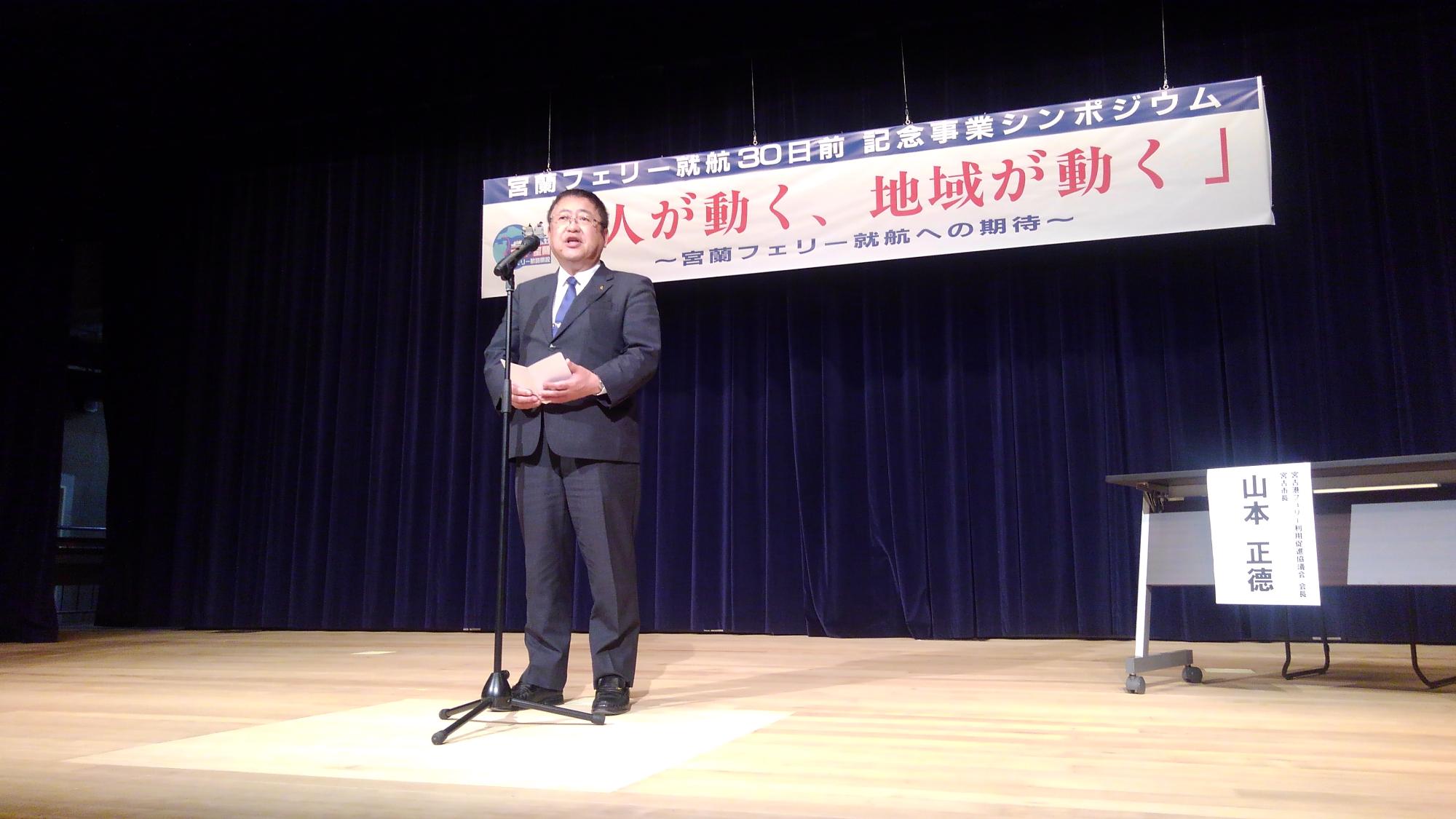 「人が動く、地域が動く」というシンポジウムの題名が書かれた横断幕が吊るされた舞台上で、スタンドマイクに向かい話をする様子の市長の写真