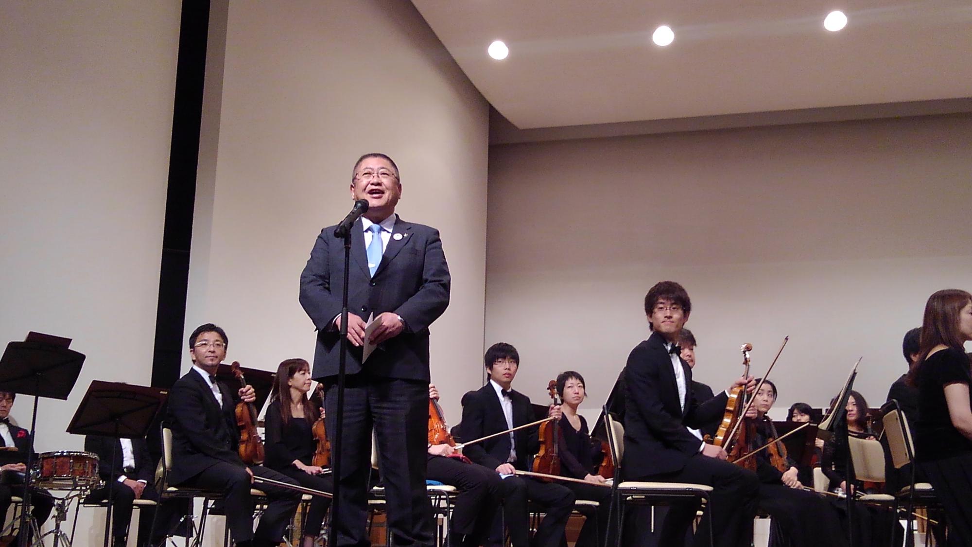 バイオリンなどの弦楽器を持ち椅子に座る交響楽団の方々の前で、スタンドマイクに向かい話をする様子の市長の写真