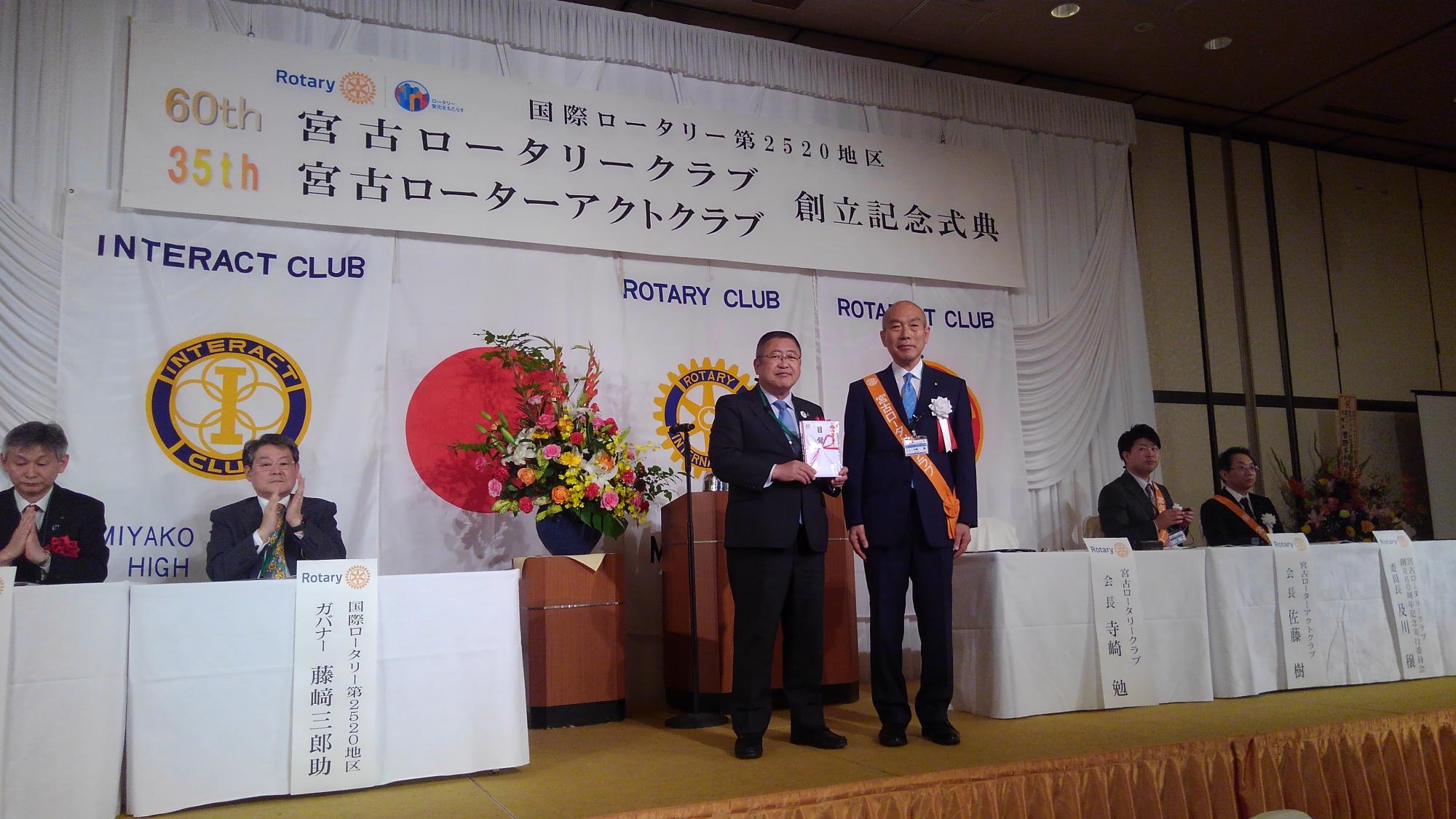 「60th 宮古ロータリークラブ 35th宮古ローターアクトクラブ 創立記念式典」と書かれたパネルが吊るされた舞台上で、目録を持つ市長と胸章を着けた男性が中央で並び、左右に並べられたテーブルの席に着き拍手をしている関係者の方々の写真
