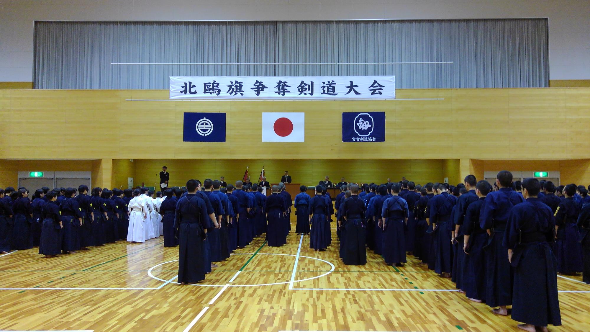 「北鷗旗争奪剣道大会」と書かれた横断幕と日本国旗、宮古市の市町村旗、宮古剣道協会の旗が吊るされた会場で、演台で話す市長と、剣道着を着て整列する大勢の参加者たちが並んでいる写真