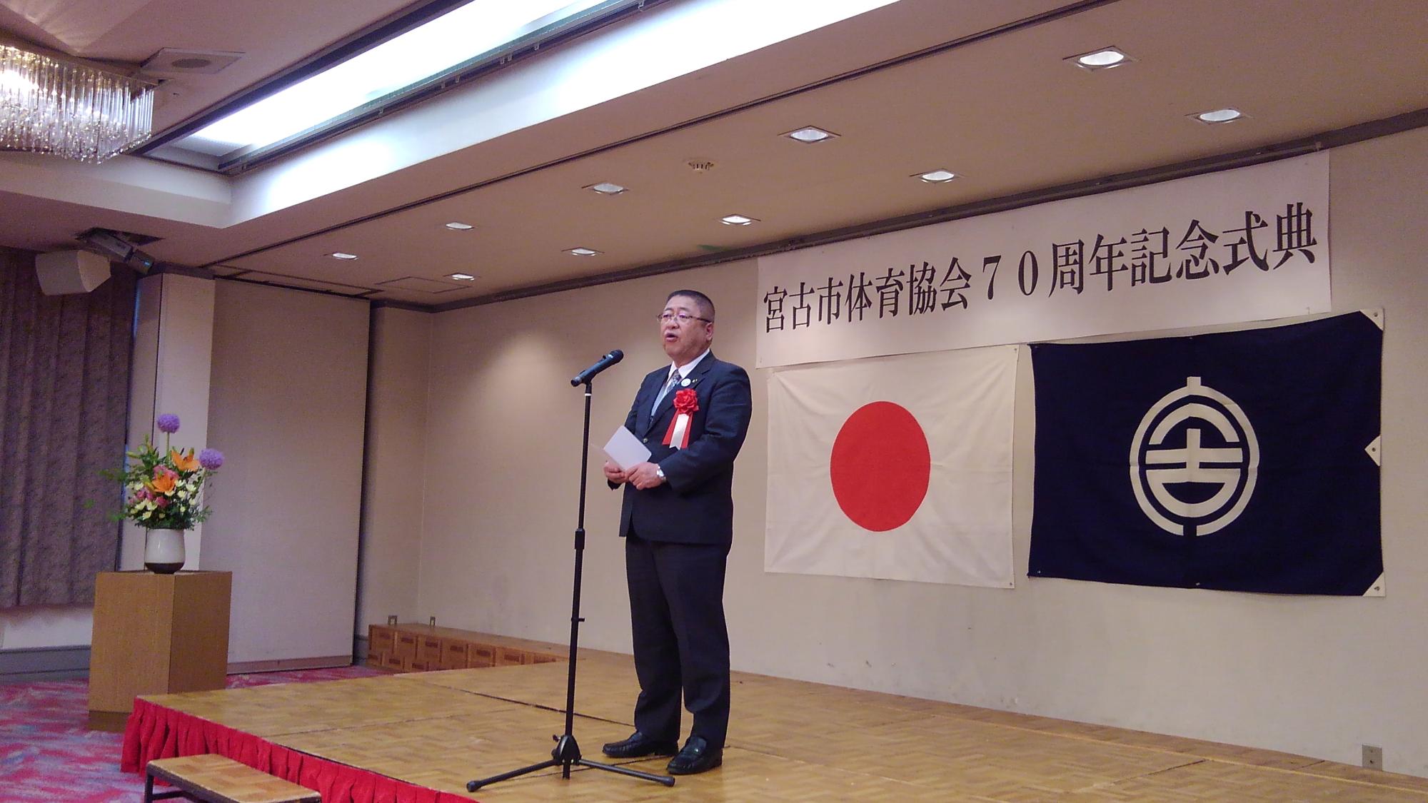 「宮古市体育協会70周年記念式典」と書かれた紙と日本国旗、宮古市の市町村旗が貼られた壁の前の舞台で、スタンドマイクに向かい話をする様子の市長の写真