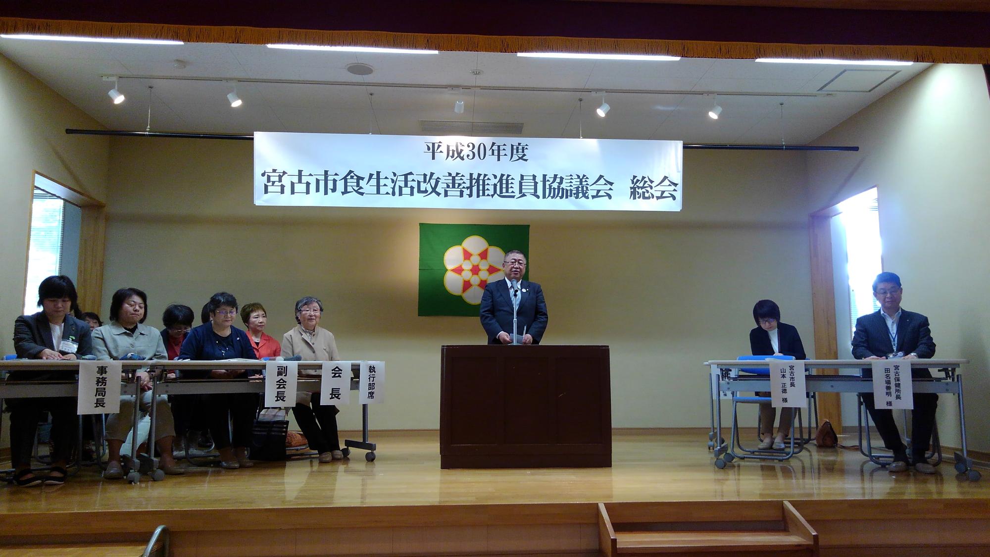 「平成30年度 宮古市食生活改善推進員協議会 総会」と書かれた横断幕の下の演台で話す市長と、演台の左右に並べられた長机の席につく関係者の方々の写真