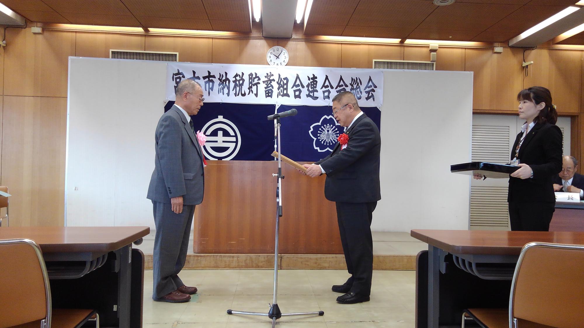 演台の前で表彰を受ける男性と、賞状を読み上げる様子の市長の写真