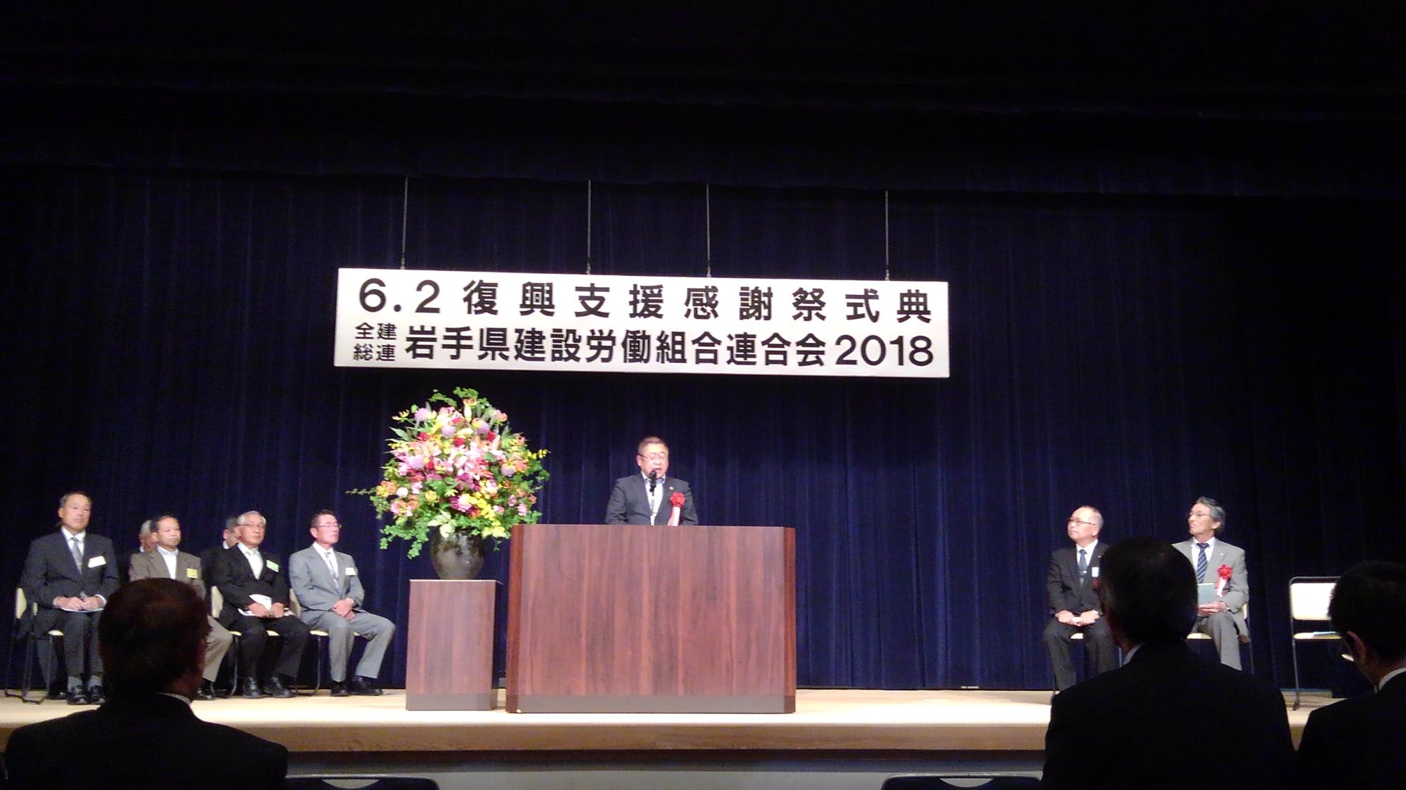 「6.2 復興支援感謝祭式典 全建総連岩手県建設労働組合連合会2018」と書かれたパネルが吊るされた舞台で、演台に置かれたマイクに向かい話をする様子の市長の写真