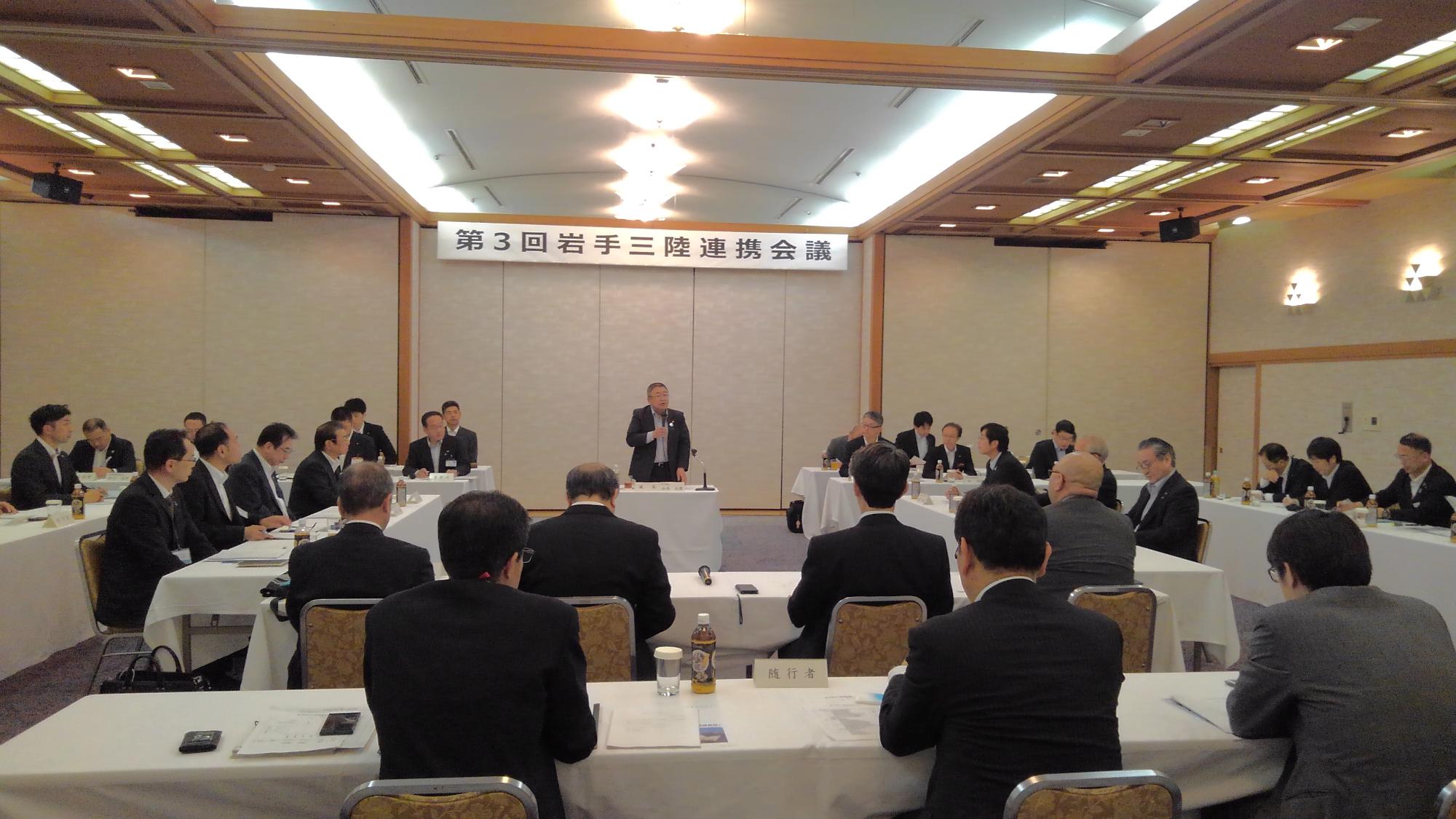 「第3回岩手三陸連携会議」と書かれた横断幕が吊るされた壁の前で立って話す市長と、コの字型に並べられたテーブルの席に着く関係者の方々の写真