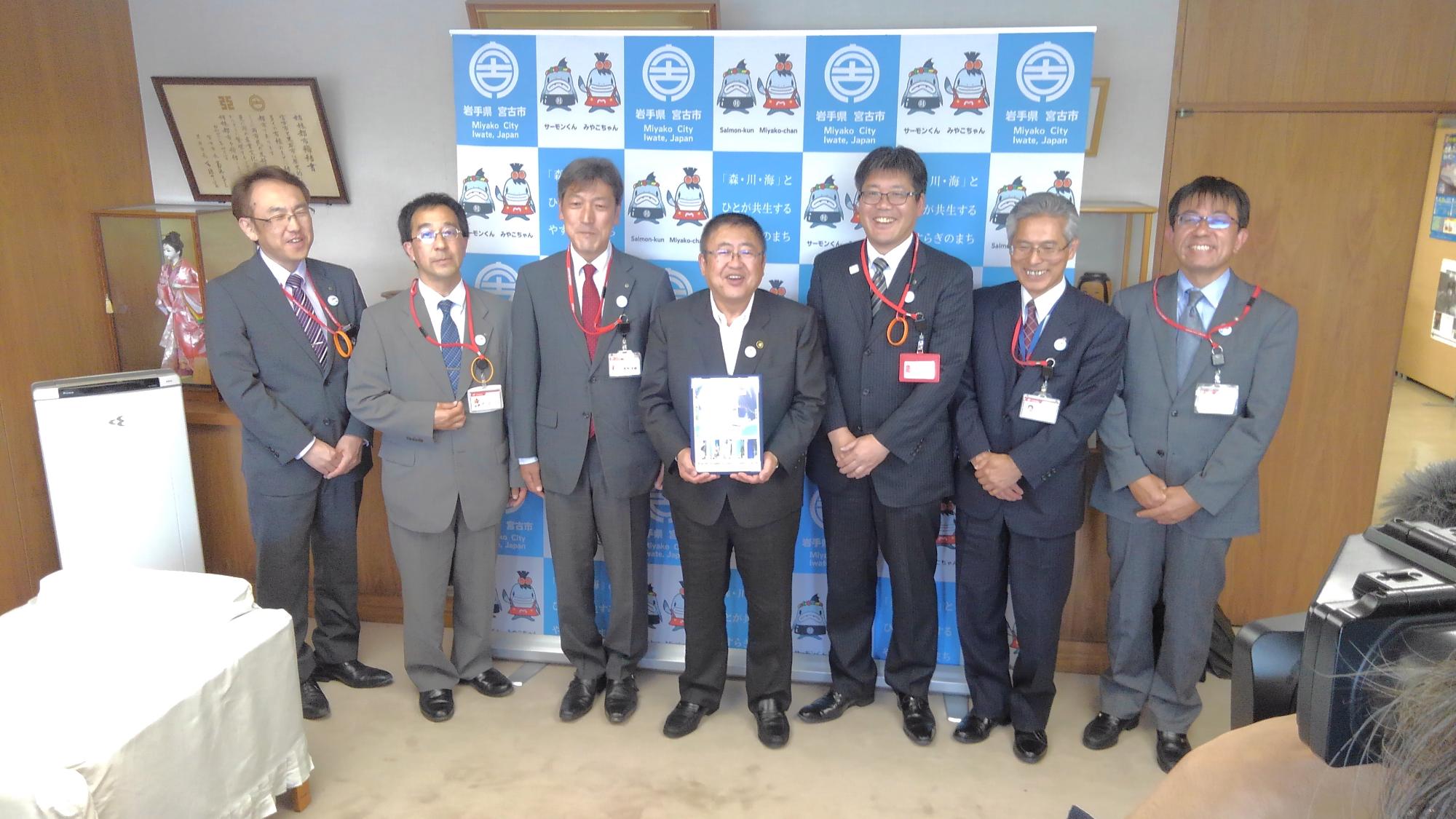 バックパネルの前で記念切手を持つ市長を中心に、関係者の男性6名が横一列に並ぶ記念写真