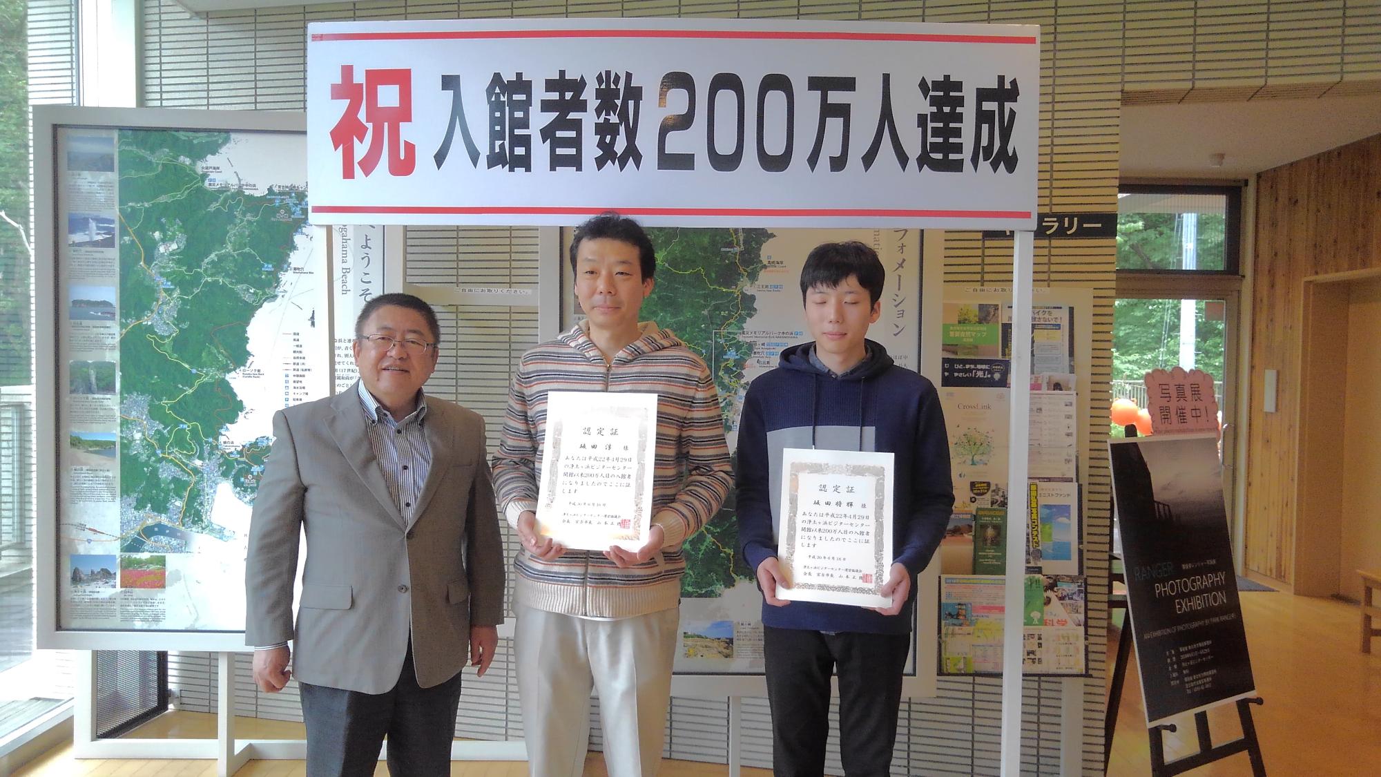 「祝 入館数200万人達成」と書かれたパネルの下で、賞状を持つ親子と市長が並んで立つ記念写真