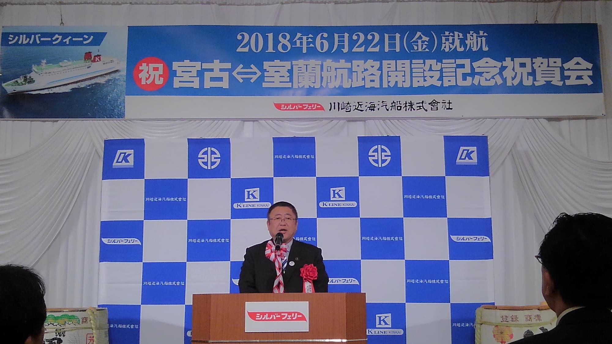 「2018年6月22日（金曜日）就航 祝宮古↔室蘭航路解説記念祝賀会」と書かれた横断幕が掛けられた壁の前で、演台に置かれたマイクに向かい話をする様子の市長の写真
